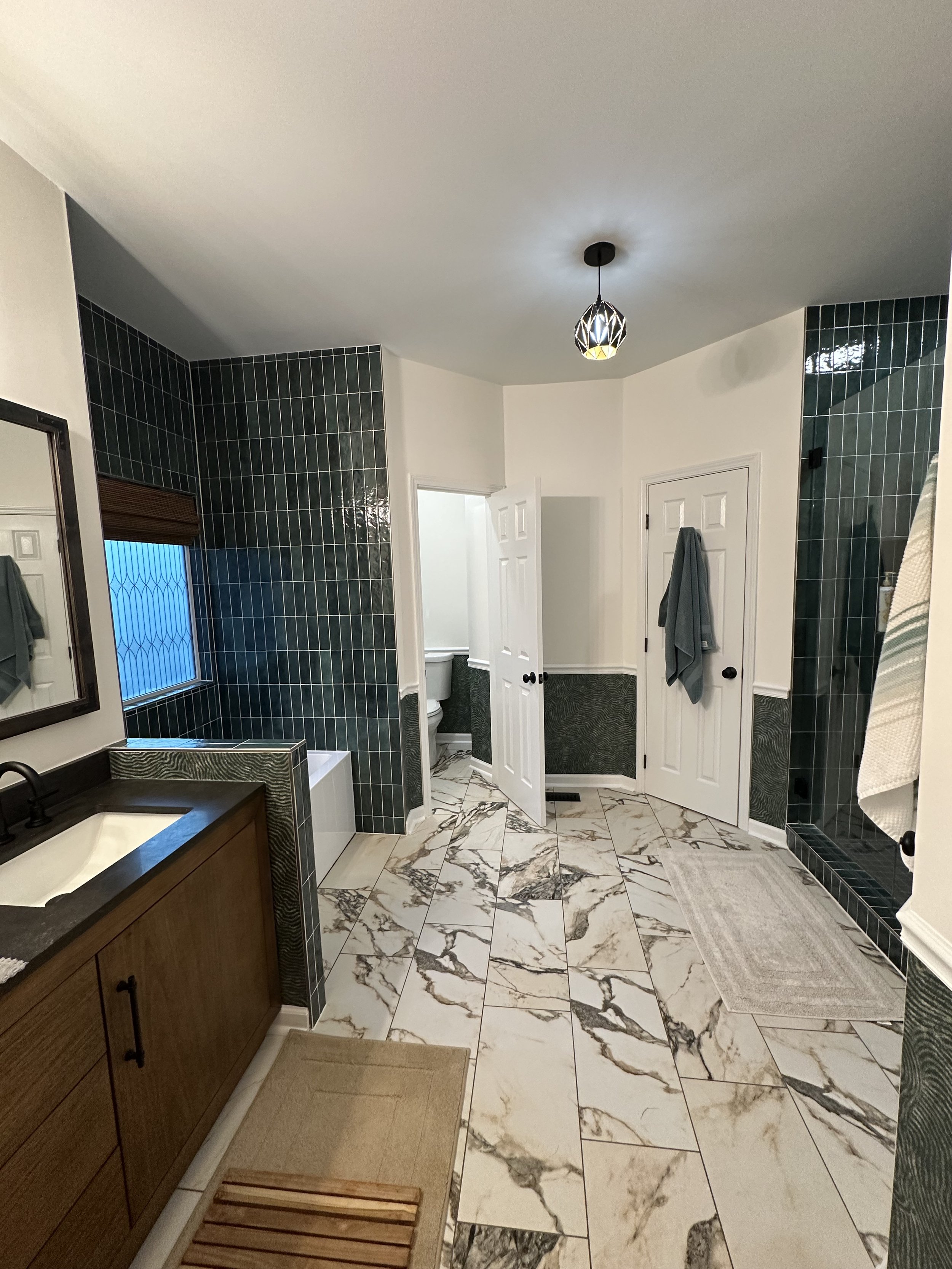 A spacious bathroom with white and dark green tiled walls, marble floor, a wooden vanity with a black countertop and sink, a mirror, a window with brown blinds, and two closed white doors, one with a towel hanging on it. There is a light fixture on t