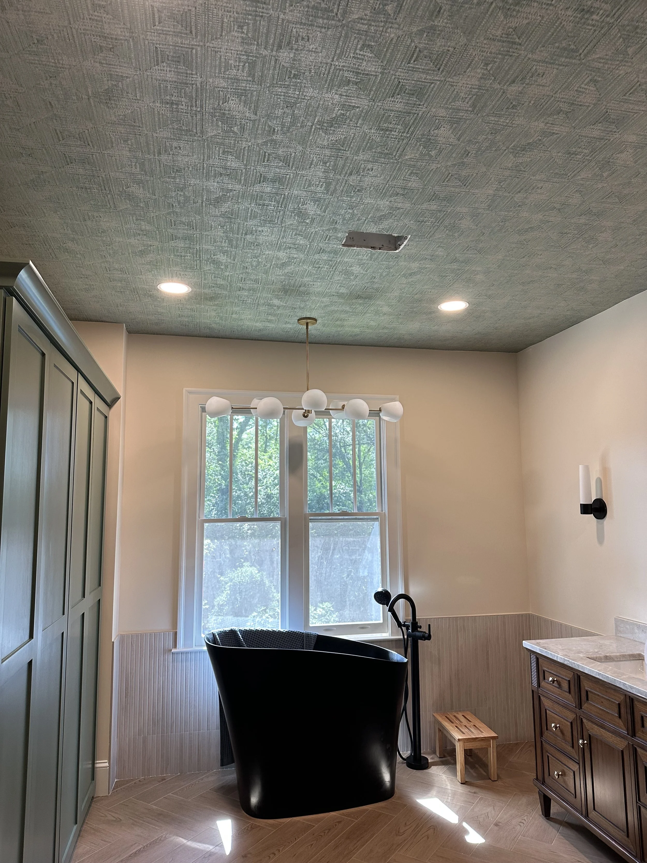 A modern bathroom with a freestanding black bathtub near a large window, wooden vanity, and a ceiling light fixture with six globe bulbs.