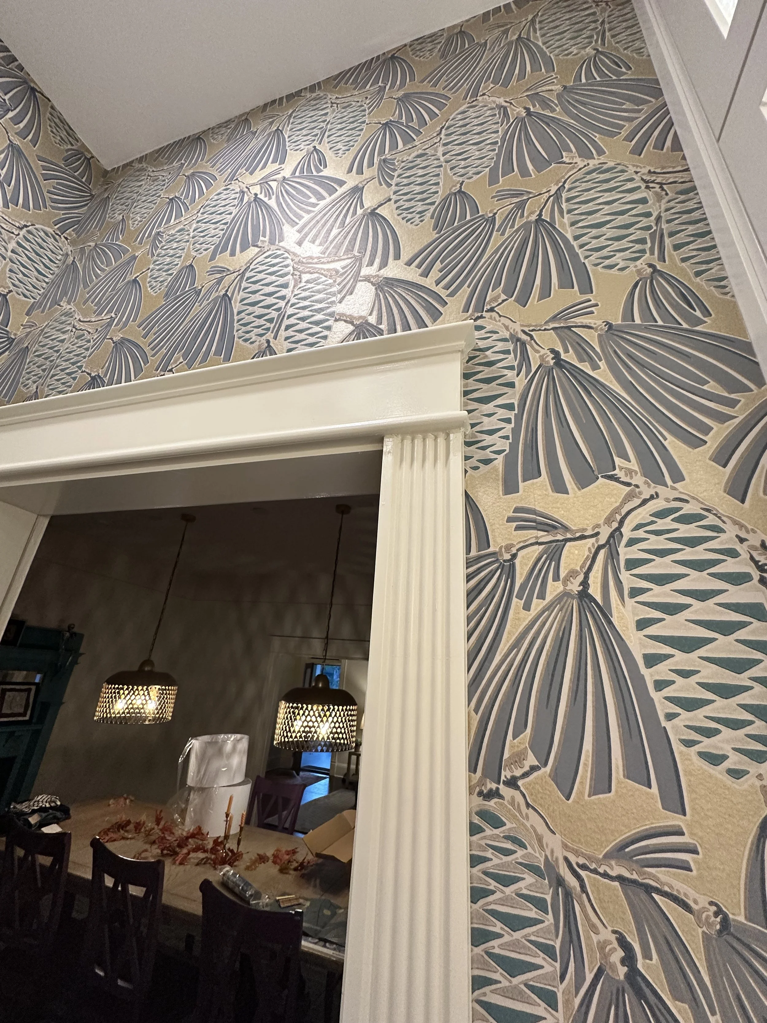 Close-up of a corner with wallpaper of large fan-shaped palm leaves in shades of blue and beige, white molding, and a doorway leading to a dining room with hanging pendant lights and a wooden table.