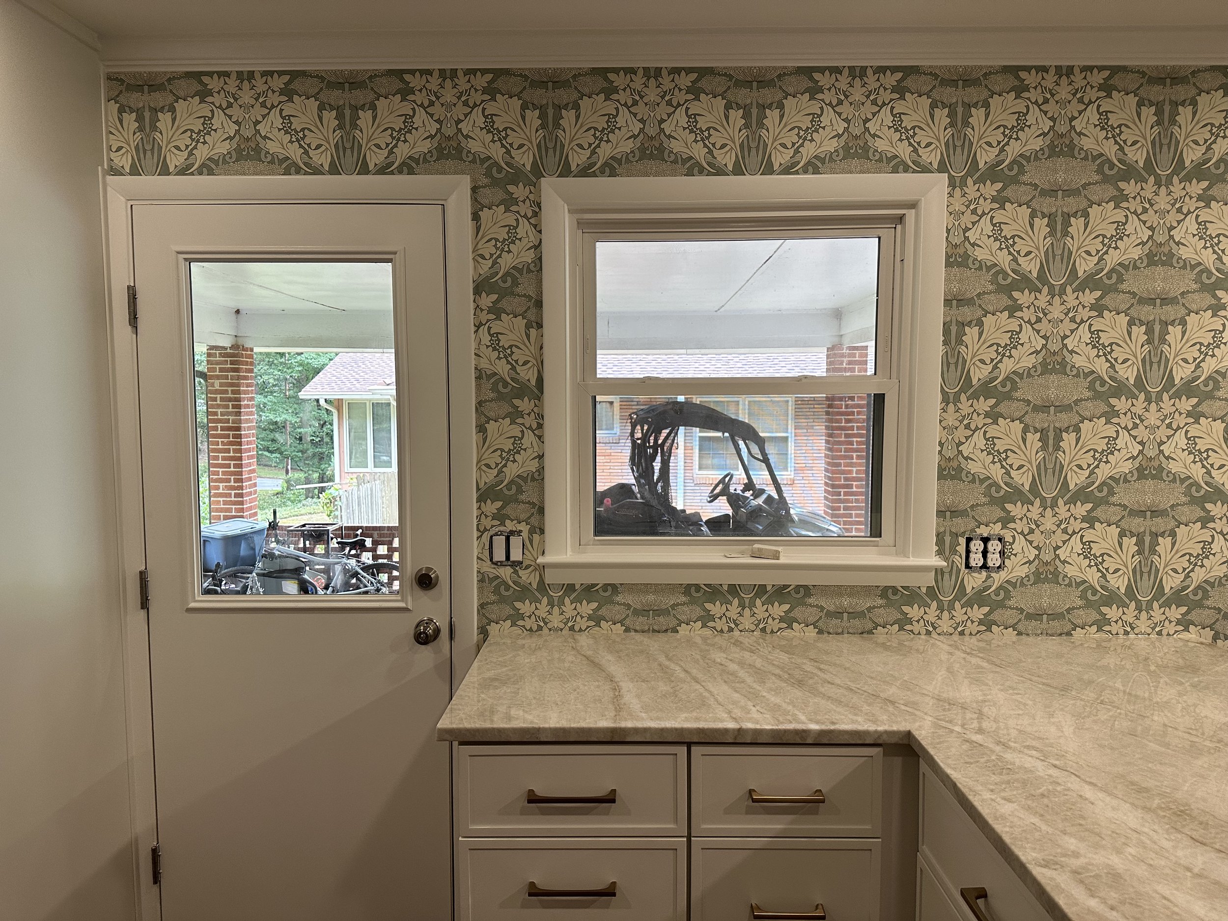 Kitchen with patterned wallpaper, white cabinetry, a marble countertop, a window, and a door leading outside, with a motorcycle visible through the window.