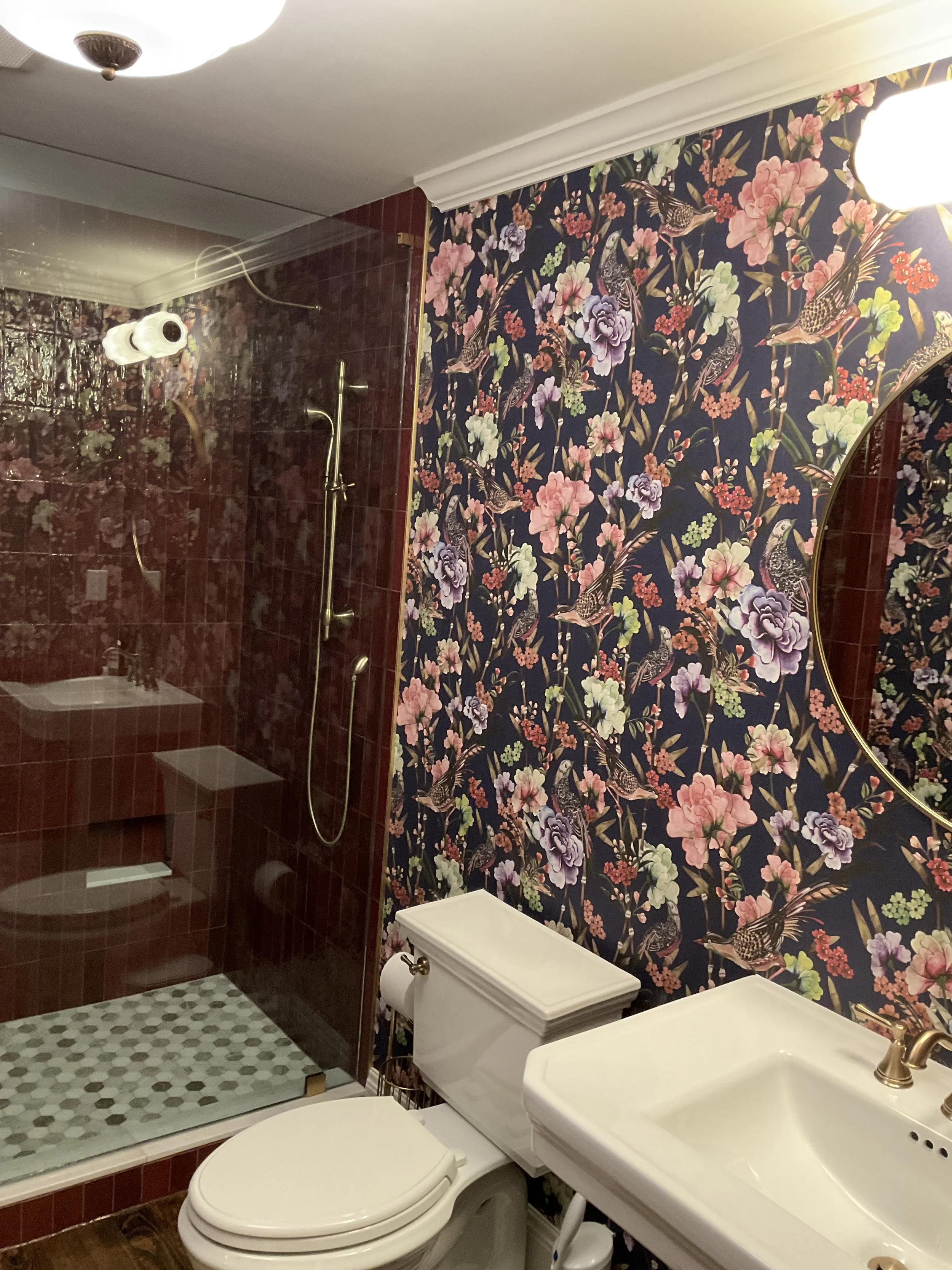 A bathroom with a floral wallpaper featuring birds, a white toilet, a white sink, a large round mirror, and a glass shower enclosure with red tiles and brass fixtures.