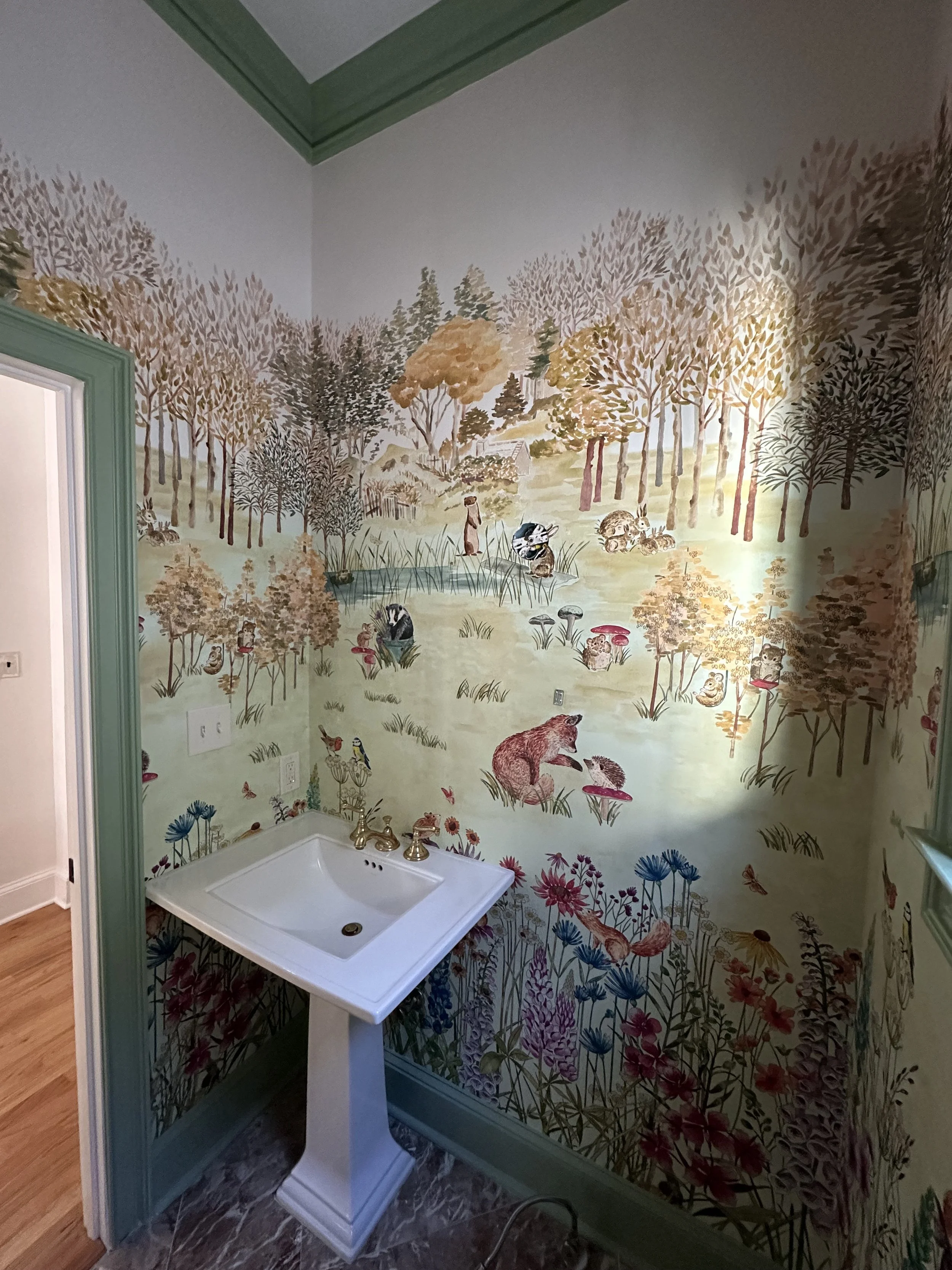A small room with painted wallpaper depicting a whimsical forest scene with various animals, mushrooms, trees, and wildflowers. There is a white pedestal sink with gold fixtures in the corner.