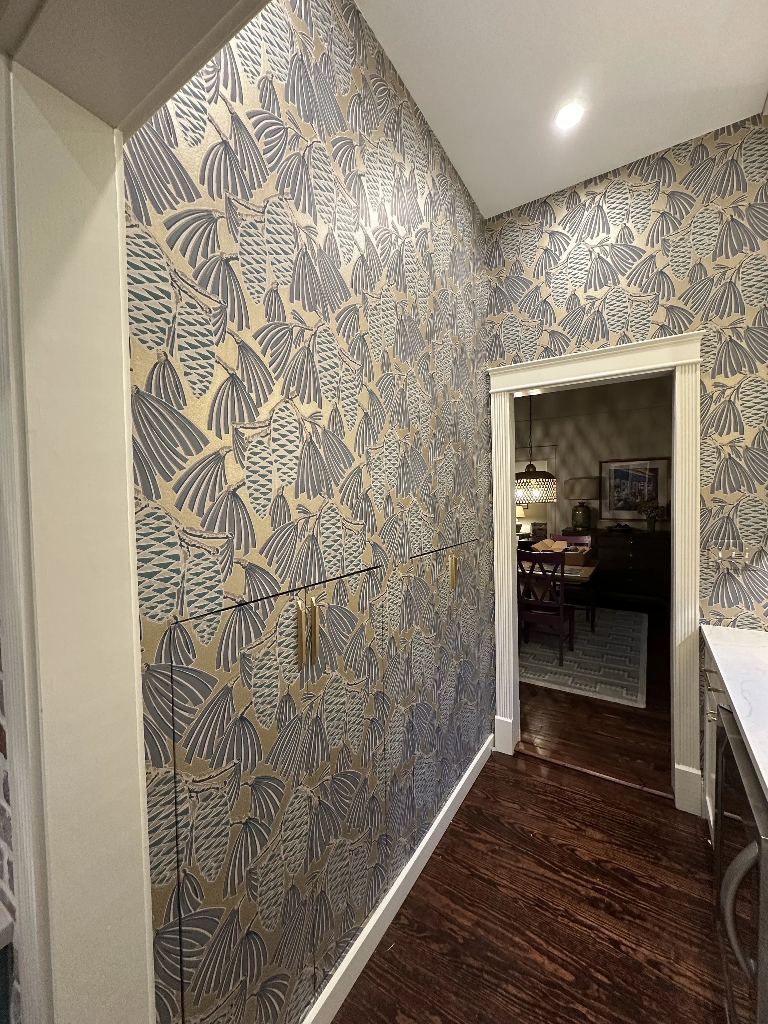 Interior view of a room with floral patterned wallpaper, a doorway leading to a dining area with a table, chairs, and a hanging light fixture, and a hardwood floor.