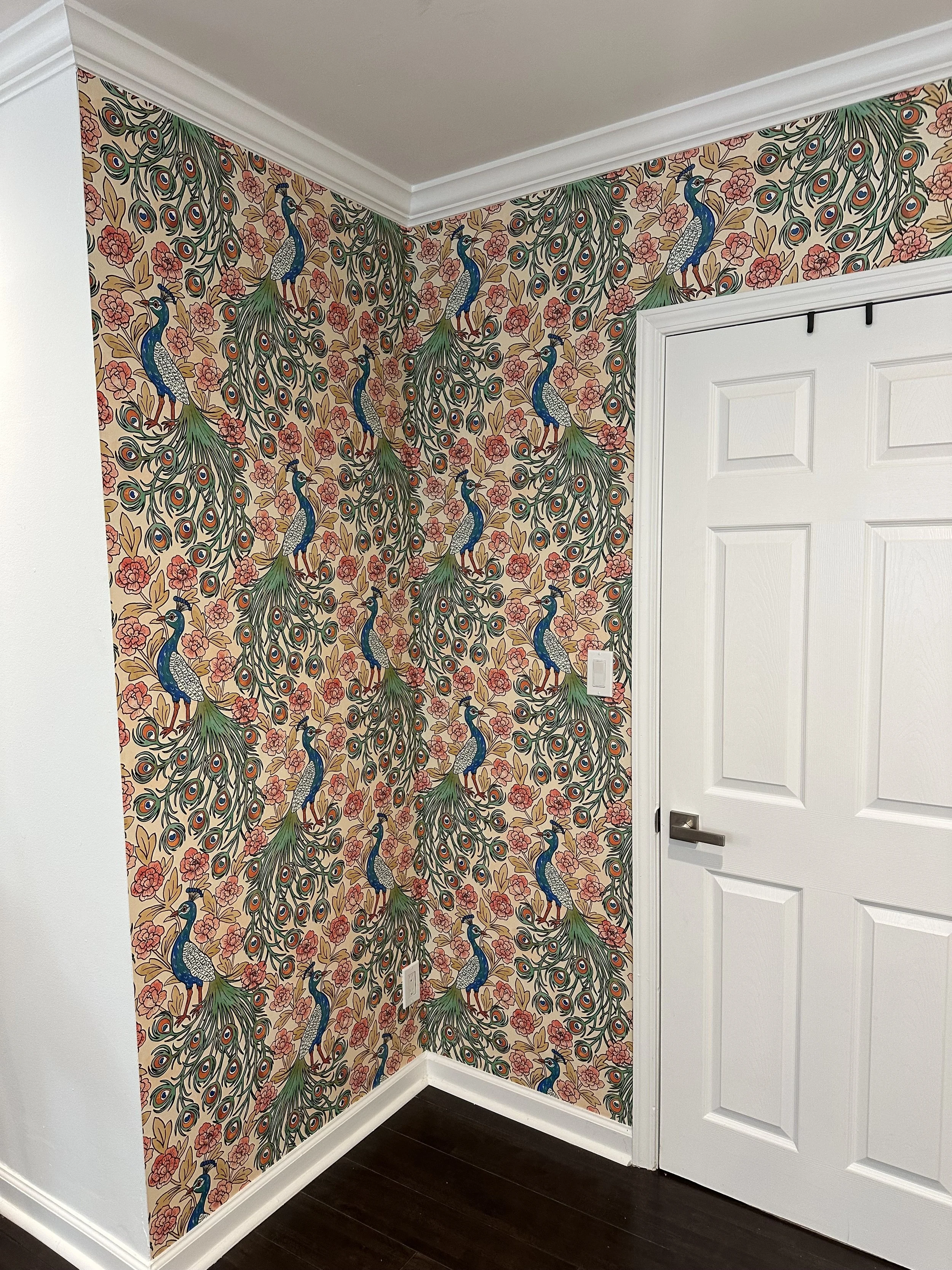 Decorative wallpaper with peacock and floral pattern in a corner of a room. The wallpaper features multiple peacocks with blue bodies, green tail feathers, and intricate eye-like patterns, surrounded by pink flowers and large green leaves. There is a