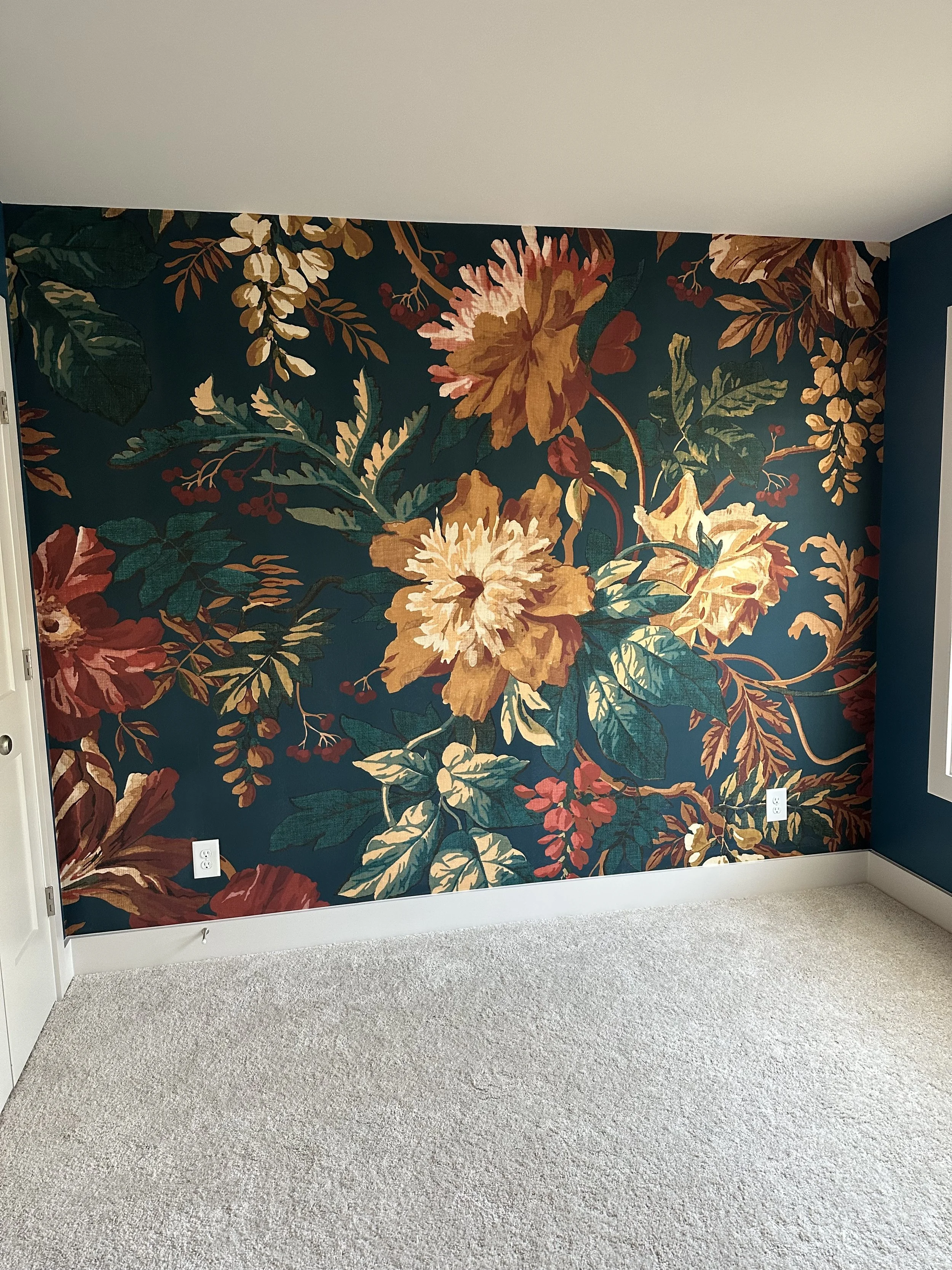 A room with a dark teal floral wallpaper featuring large yellow, orange, and red flowers with green leaves. The room has beige carpet and white baseboards, with electrical outlets visible on the wall.