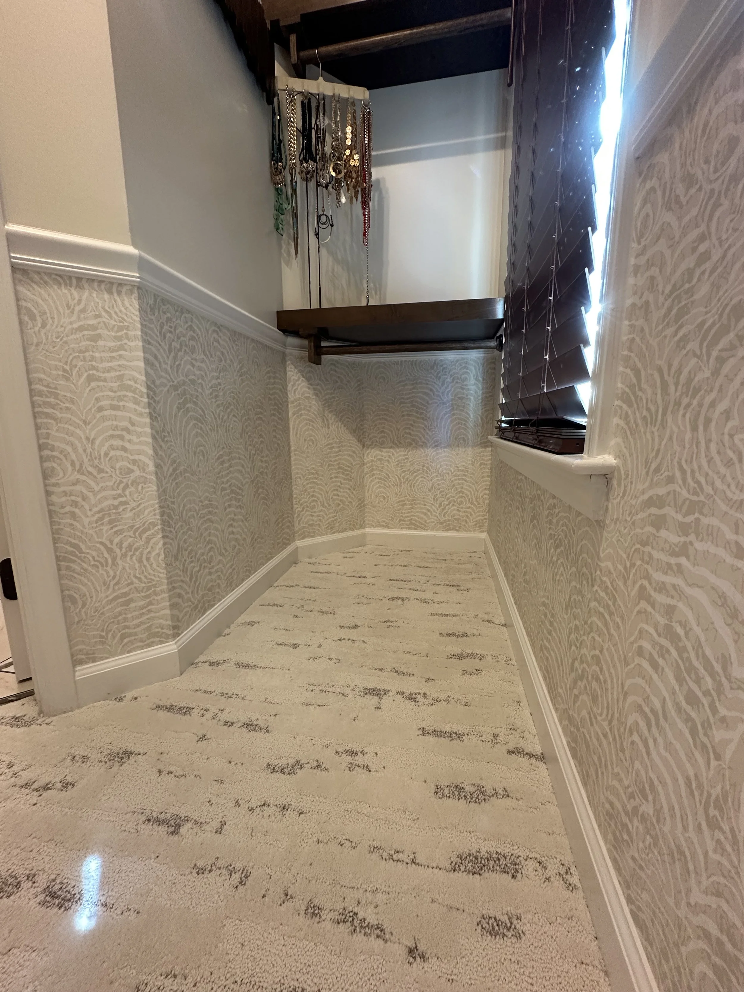 A small, narrow room with beige patterned wallpaper and a light-colored carpeted floor. There is a window with dark blinds on the right side and a built-in shelf or table on the back wall. A jewelry stand with various necklaces is visible on the left