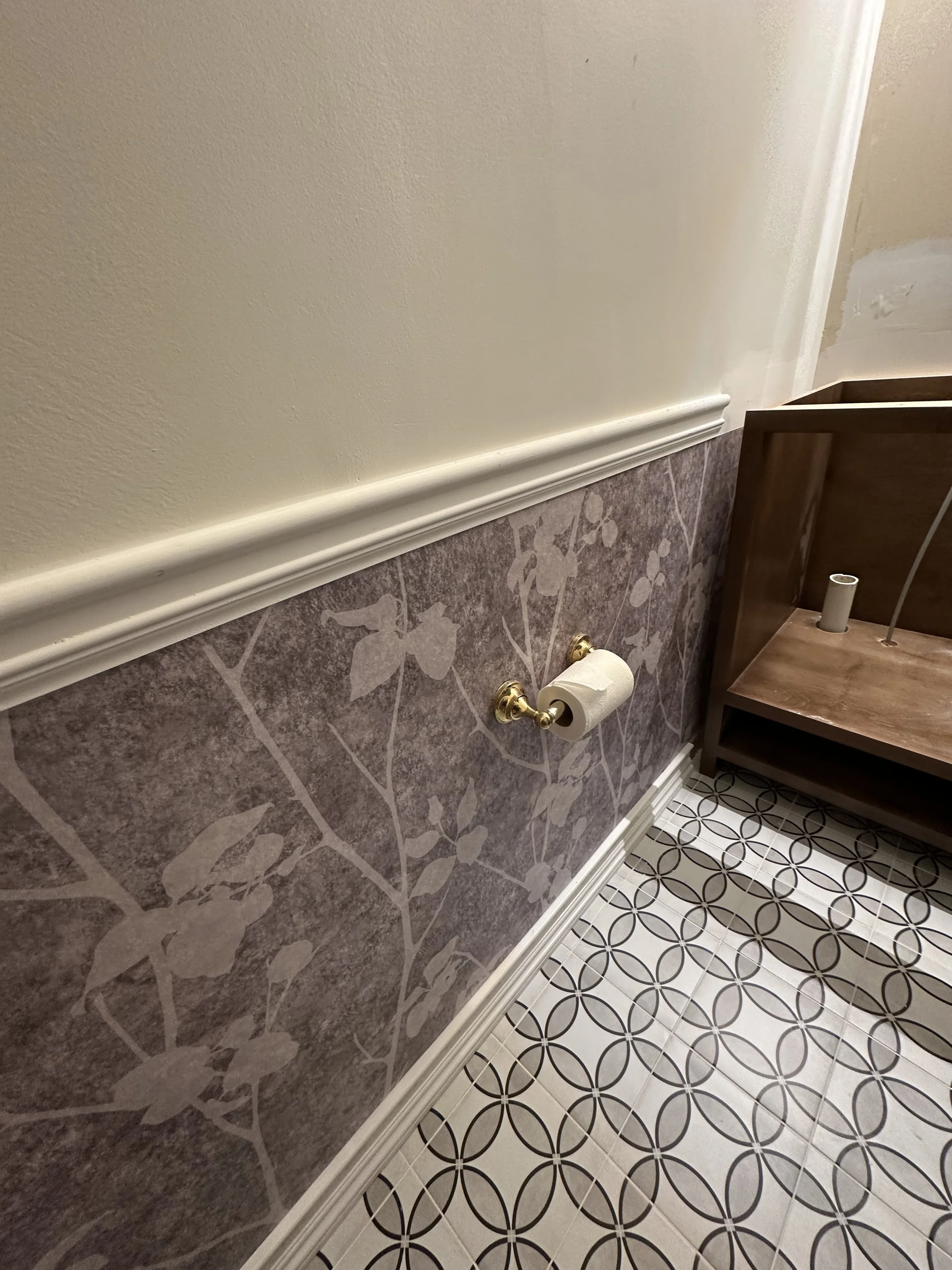 Bathroom wall with an empty toilet paper holder, a section of a patterned floor, and a wooden piece of furniture in the corner.