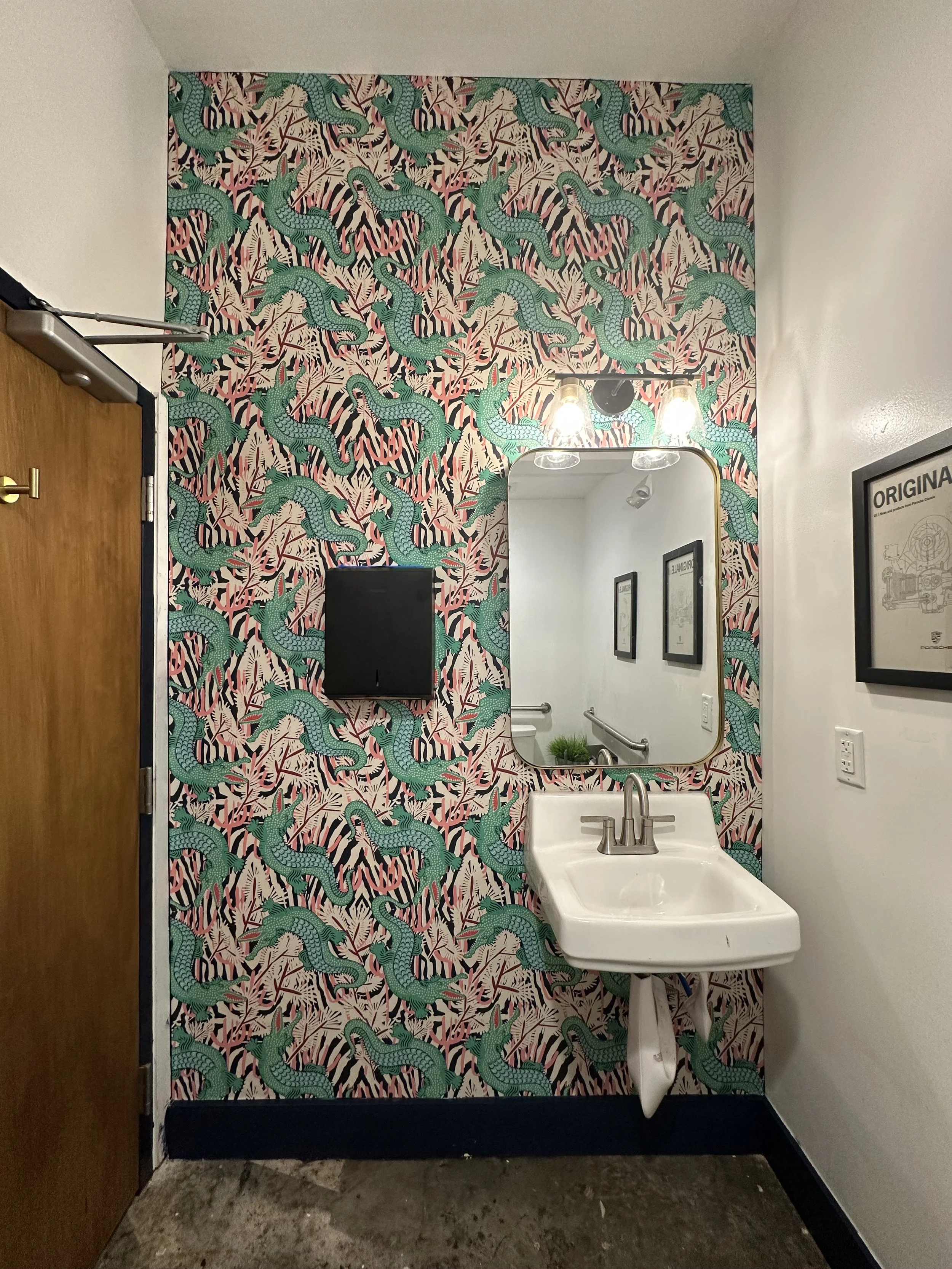 A room with a zigzag patterned wallpaper featuring green lizards and black and pink plants. There is a mirror above a white sink, with two light bulbs above the mirror. On the wall to the right, there is a framed picture. The door on the left has a b