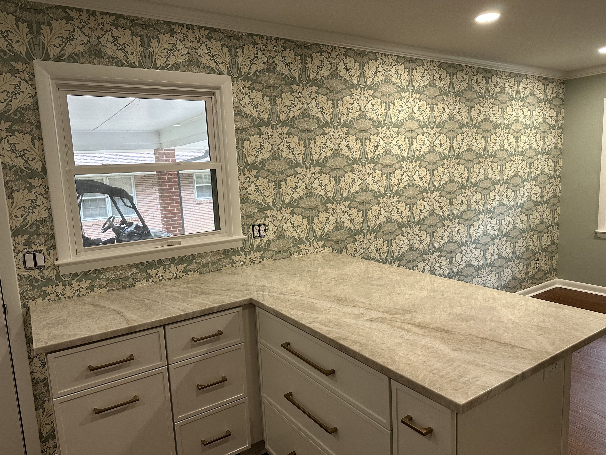 Kitchen with a large marble countertop, white cabinets with gold handles, a window with a view outside, patterned wallpaper, and recessed ceiling lights.