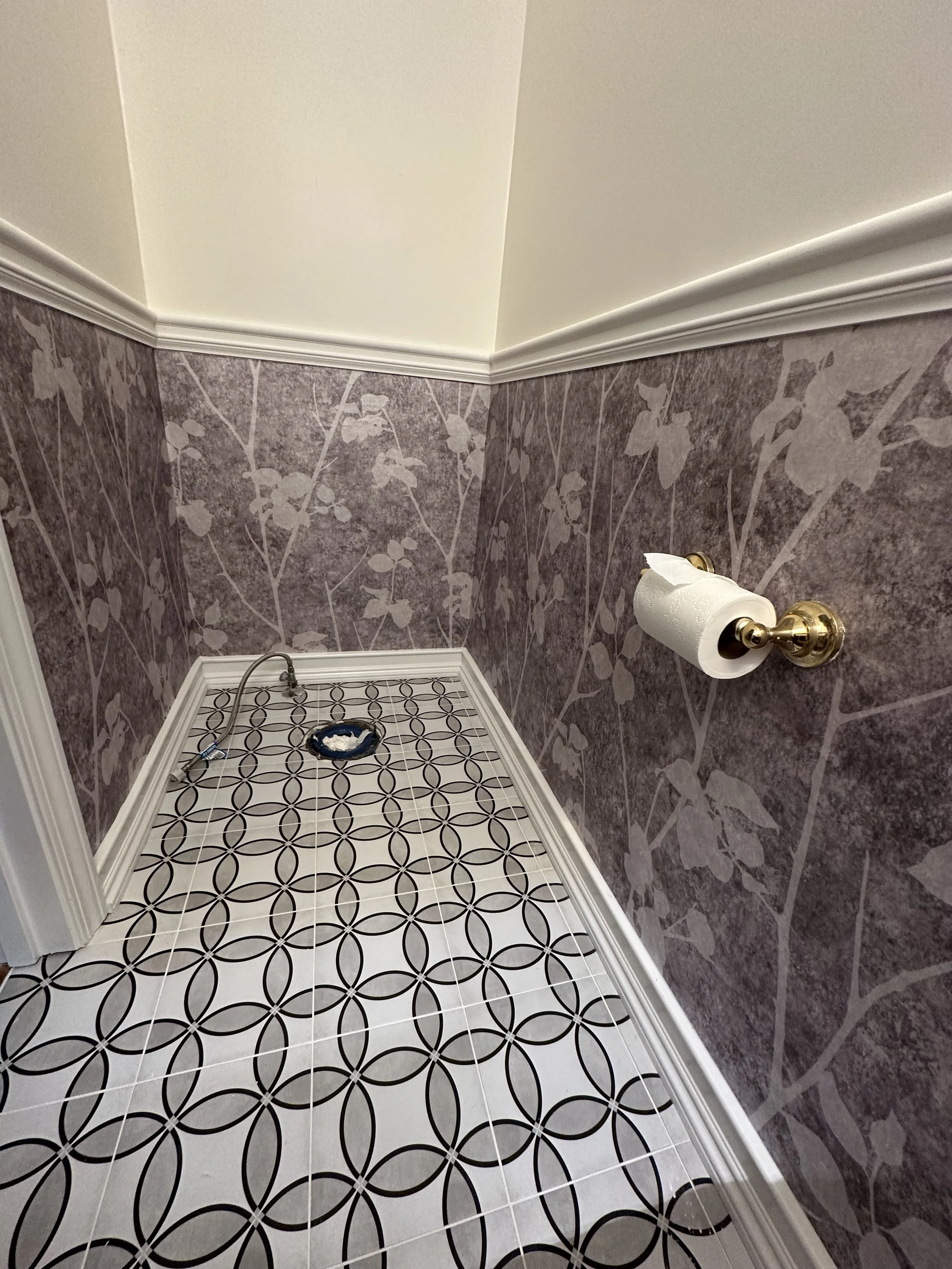 A small, empty bathroom corner with a decorative black and white tile floor, purple floral wallpapered walls, a white ceiling, white crown molding, and a brass toilet paper holder with a roll of toilet paper, with a cooking hose and blue tape on the 