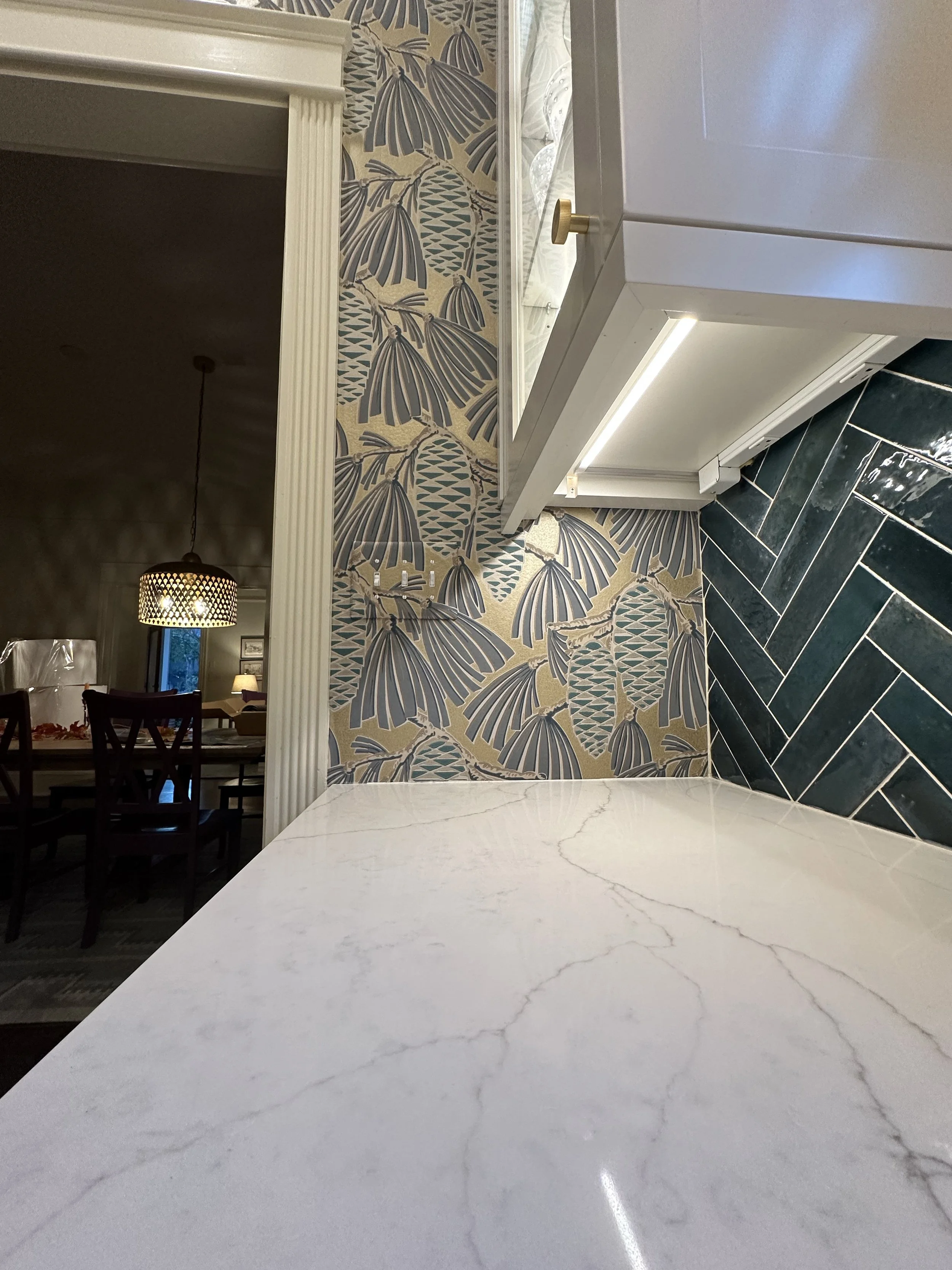 Close-up view of a kitchen countertop with white marble surface, black herringbone tile backsplash, patterned wallpaper with gold, black, and beige tropical foliage, and a white cabinet with a small gold handle.