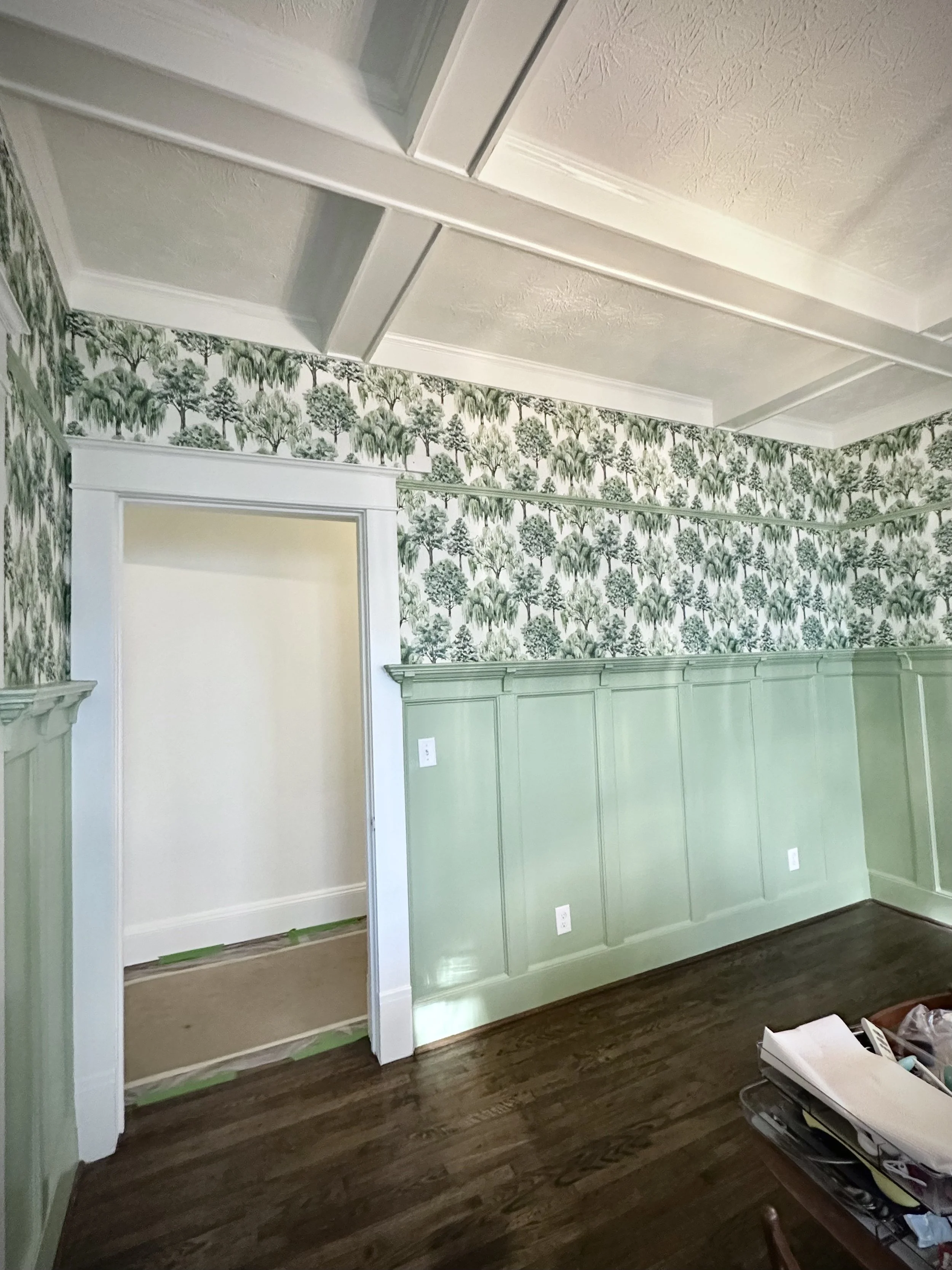 Interior of a room with green floral wallpaper, white trim, and dark hardwood flooring. A door opening leads to another room with beige walls, and construction materials are on the floor.