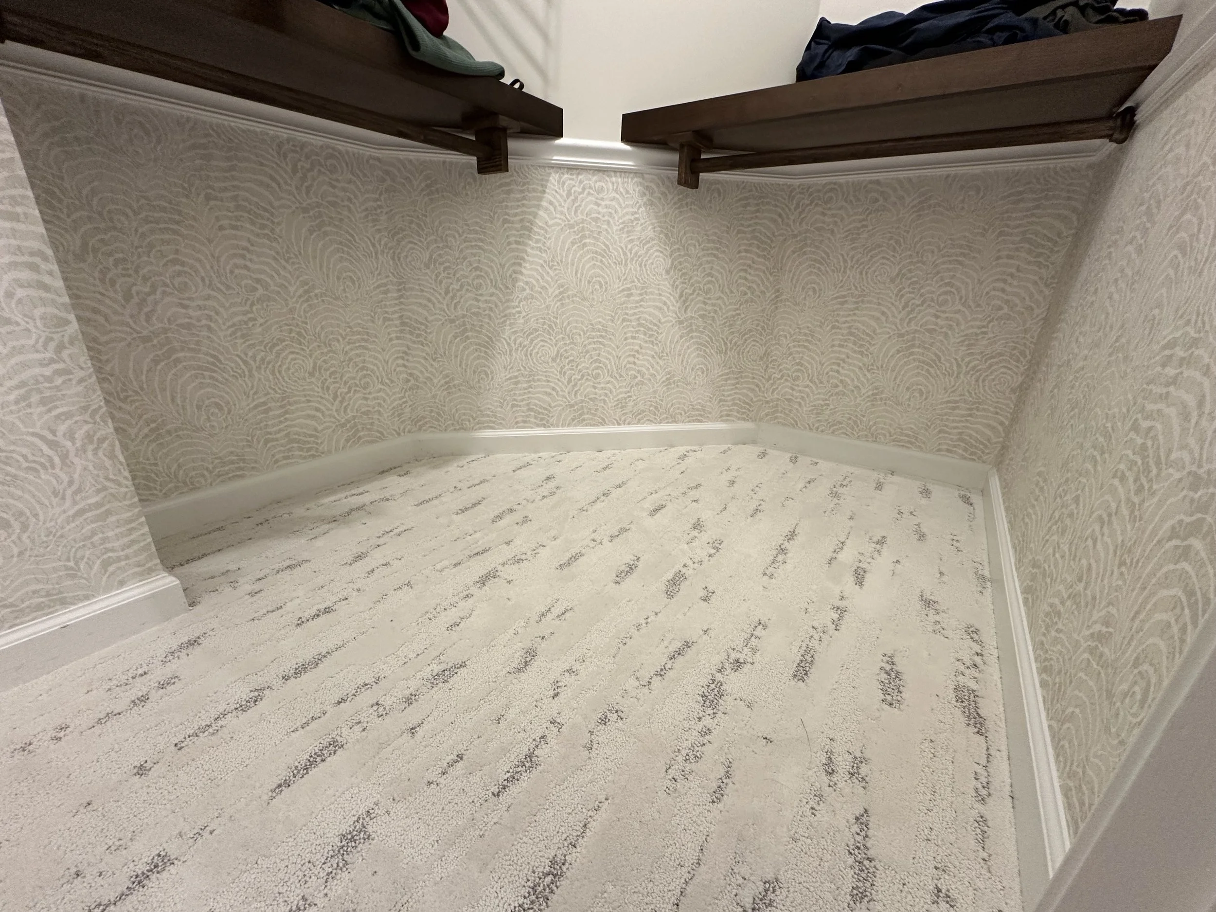 Empty walk-in closet with beige patterned wallpaper, light-colored carpet, and two dark wooden shelves on the walls.