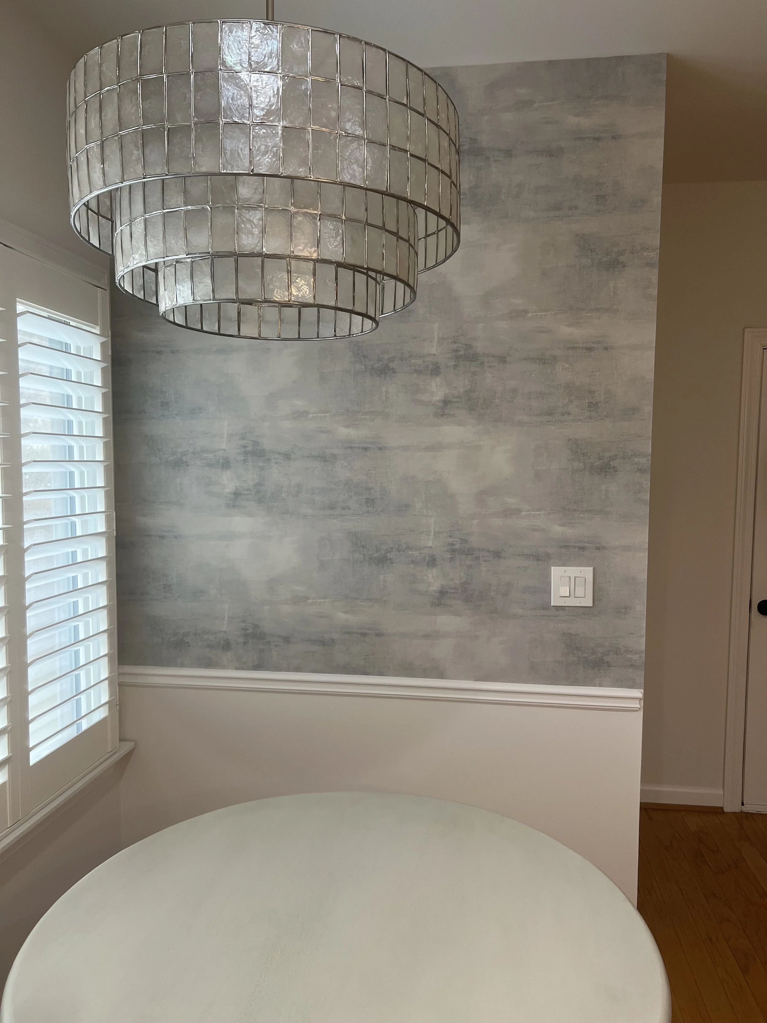 A round white table in front of a wall with a two-tone textured gray finish. A large layered pendant light with a metallic frame hangs above the table. There is a window with white plantation shutters on the left and a white electrical switch on the 