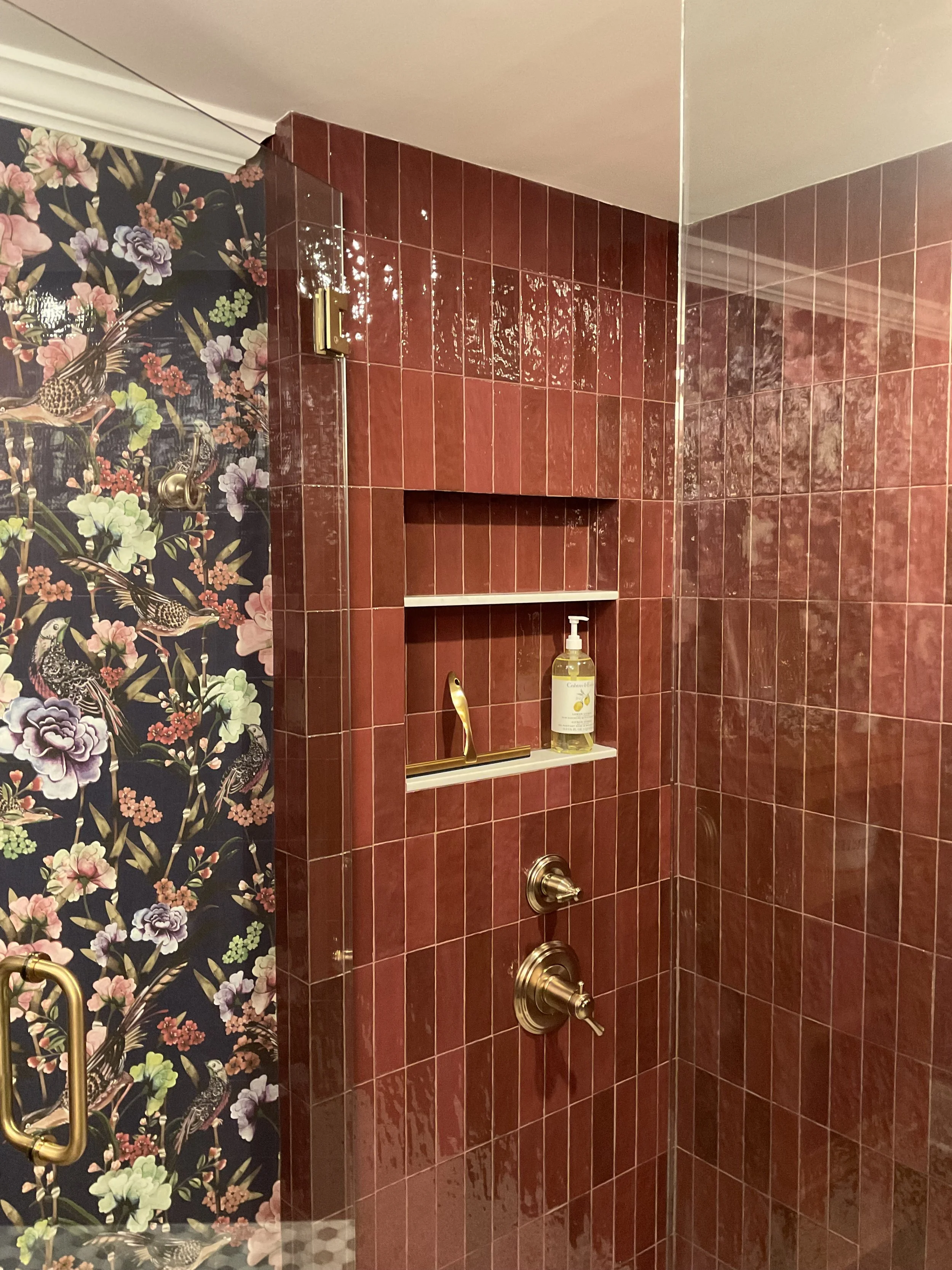 Shower area with red tiles and built-in shelves, gold fixtures, and floral-patterned wallpaper on one wall.