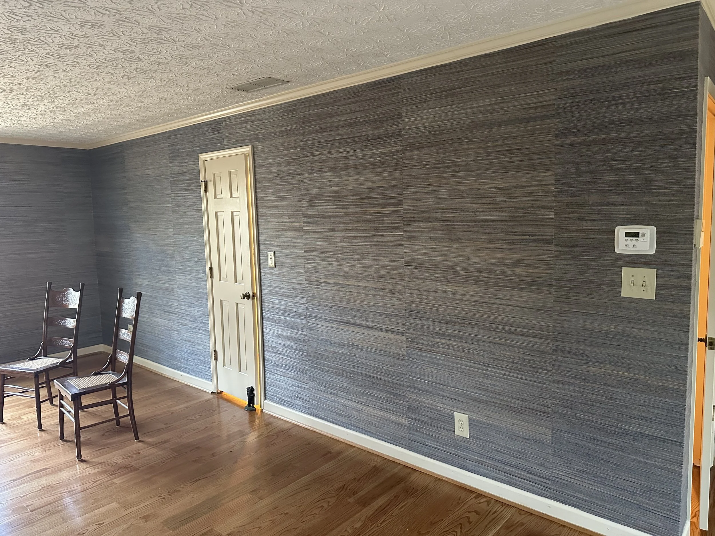 Empty dining room with grey textured wall, hardwood floor, white door, thermostat, outlet, and chairs.