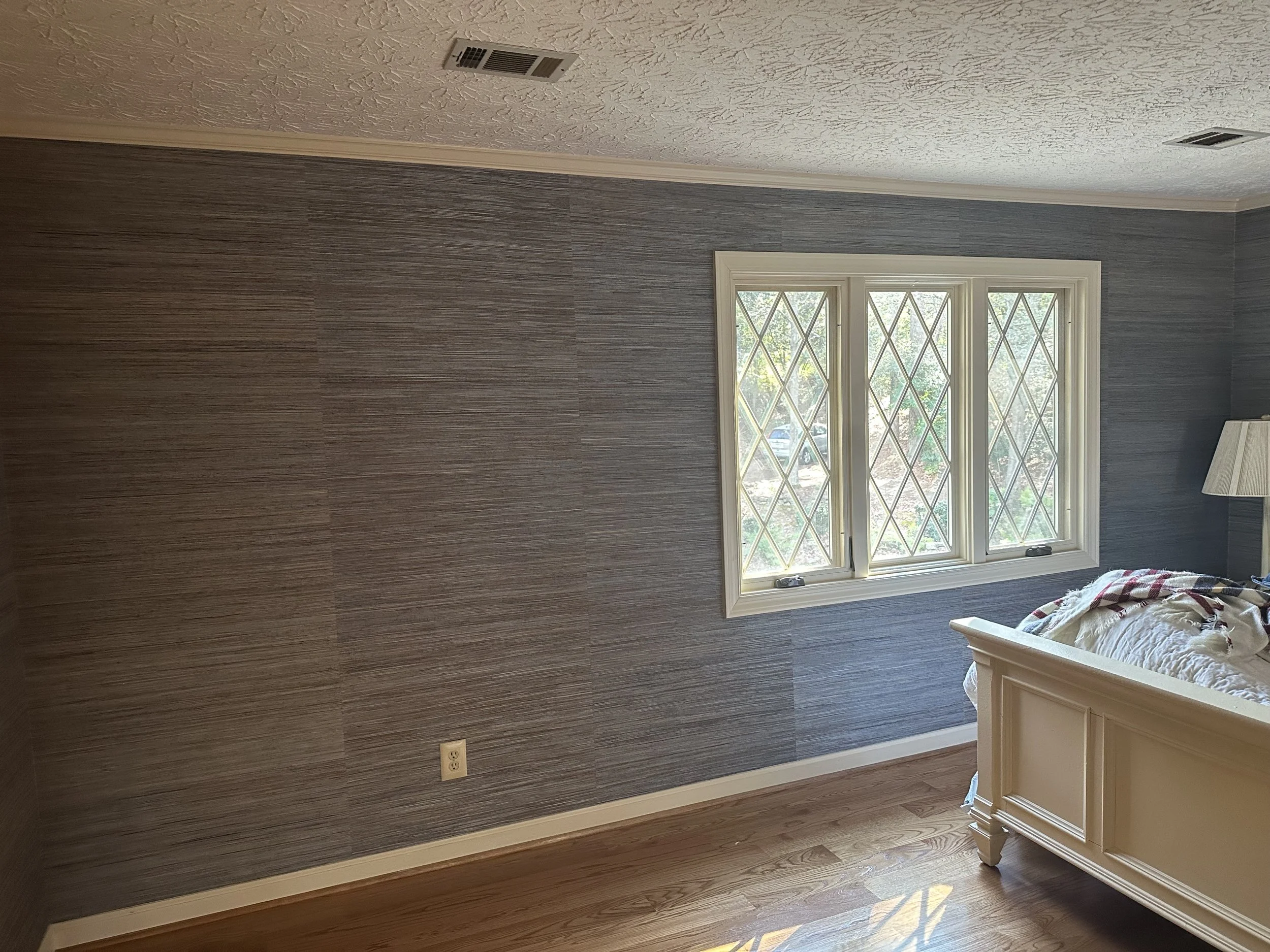 A bedroom with gray textured wallpaper, a window with decorative diamond-shaped panes, and a white bed frame partially visible on the right side. Natural light is coming through the window, illuminating the wooden floor.