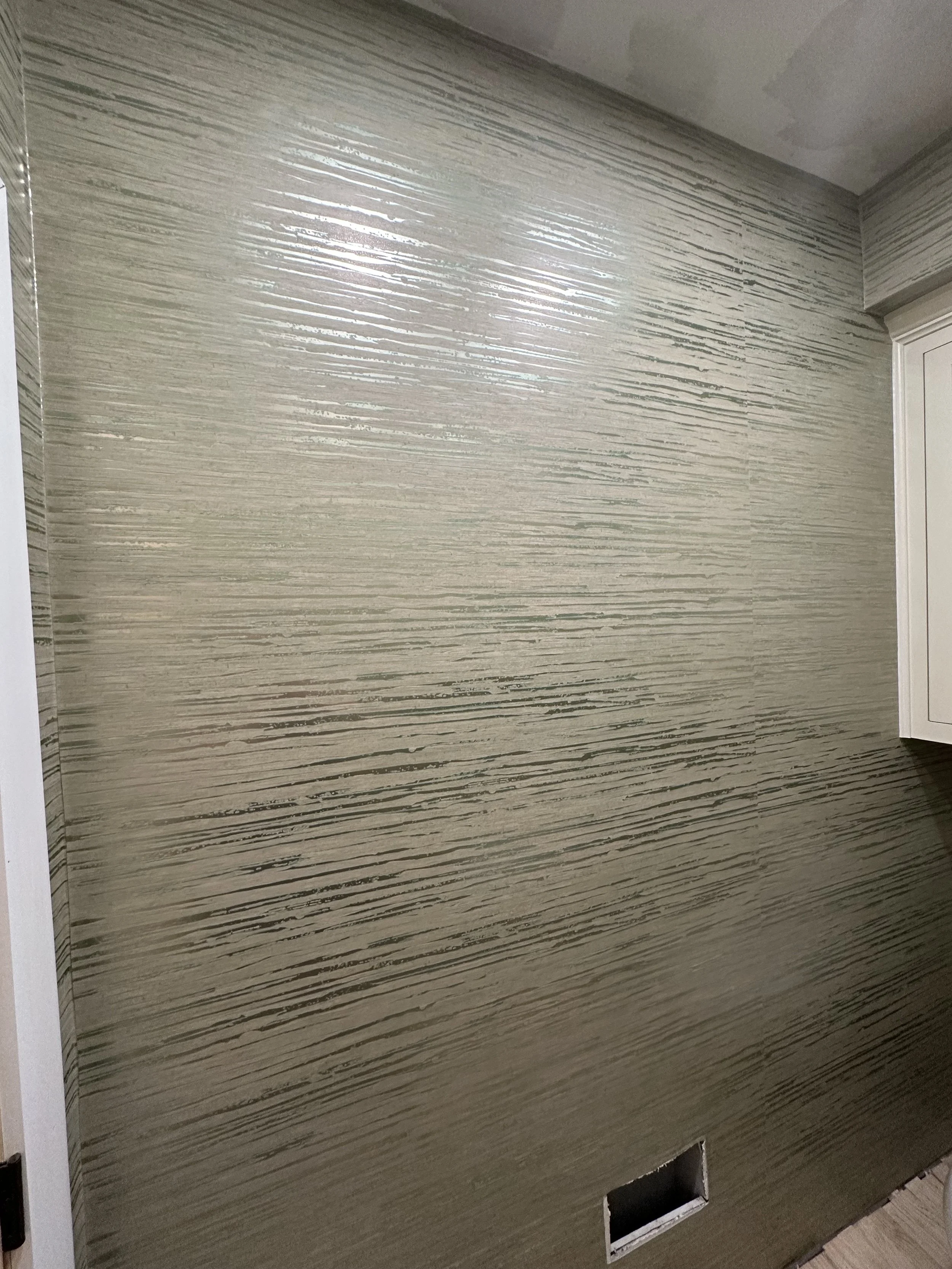 A wall with textured beige and gray horizontal streaks, with an opening near the bottom for an electrical outlet or vent, in a room under renovation.