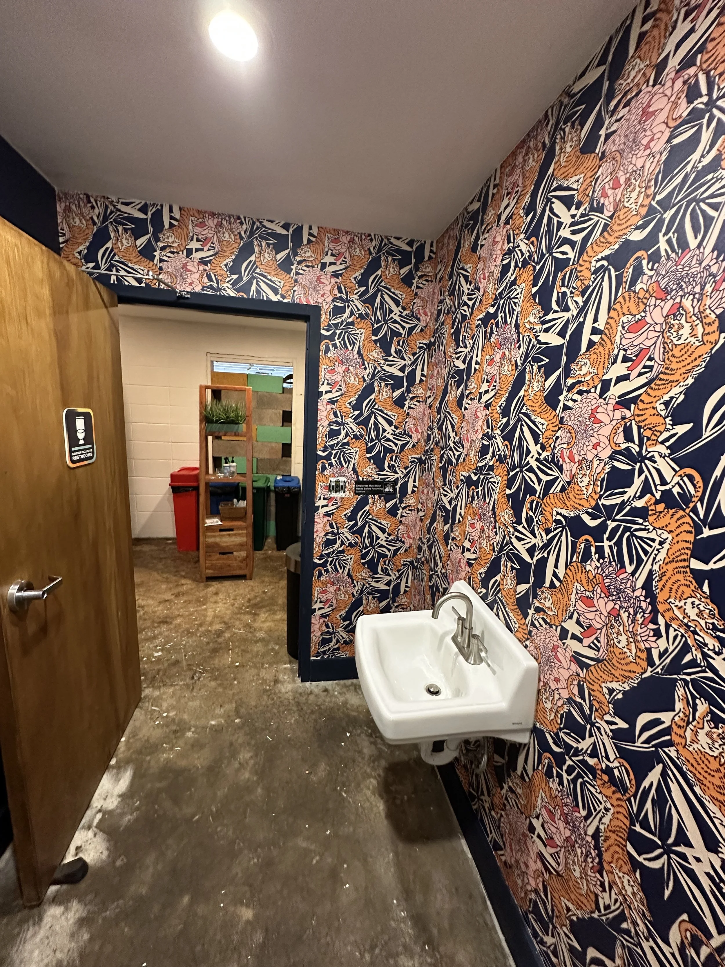 Public restroom with tiger-themed wallpaper, a small white sink, and a wooden door leading to the toilet area with trash bins and a wooden shelf in the background.
