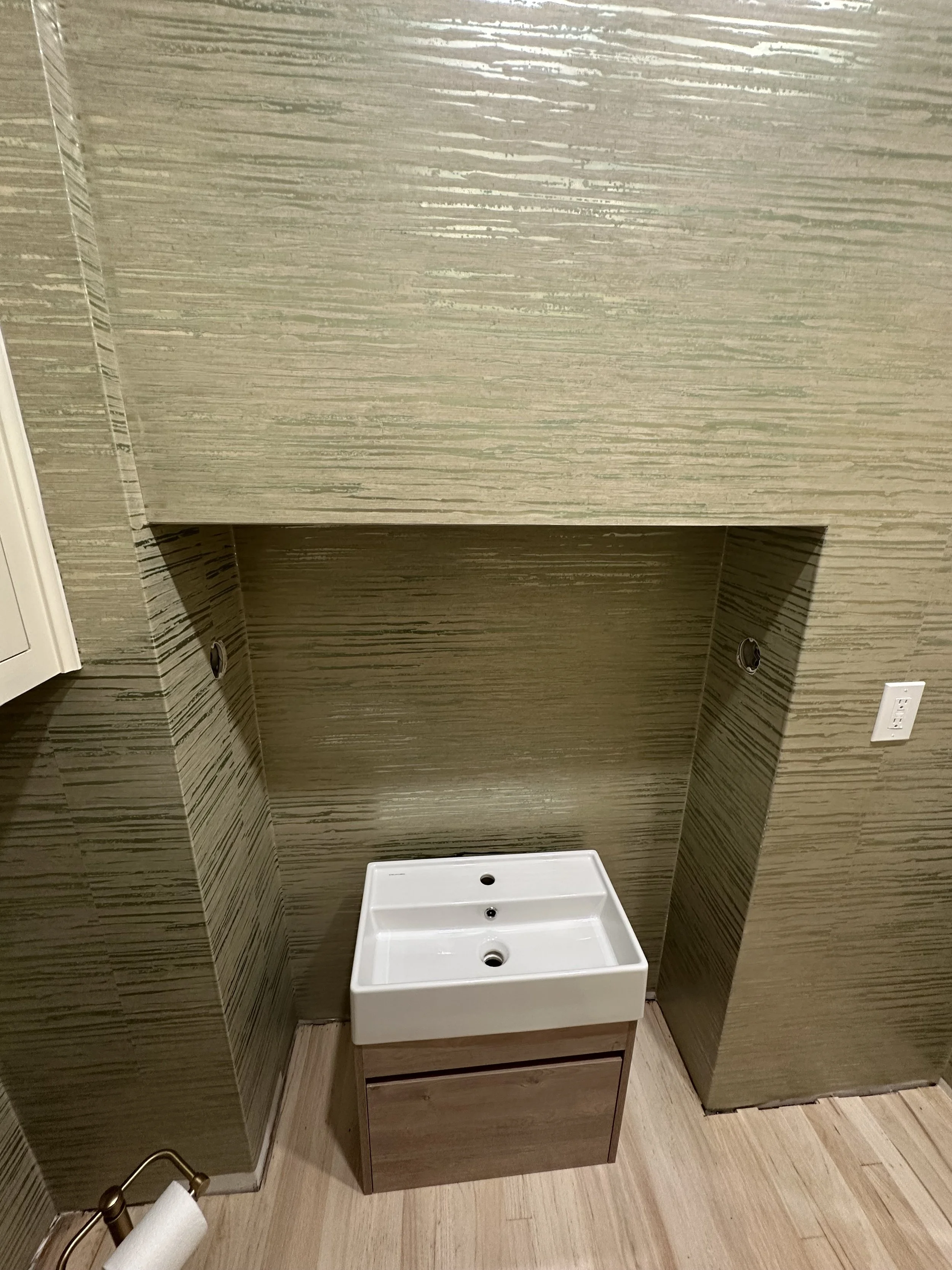 Empty bathroom space with a small white sink base cabinet against a textured greenish-brown wall, with an outlet and a paper towel holder visible.