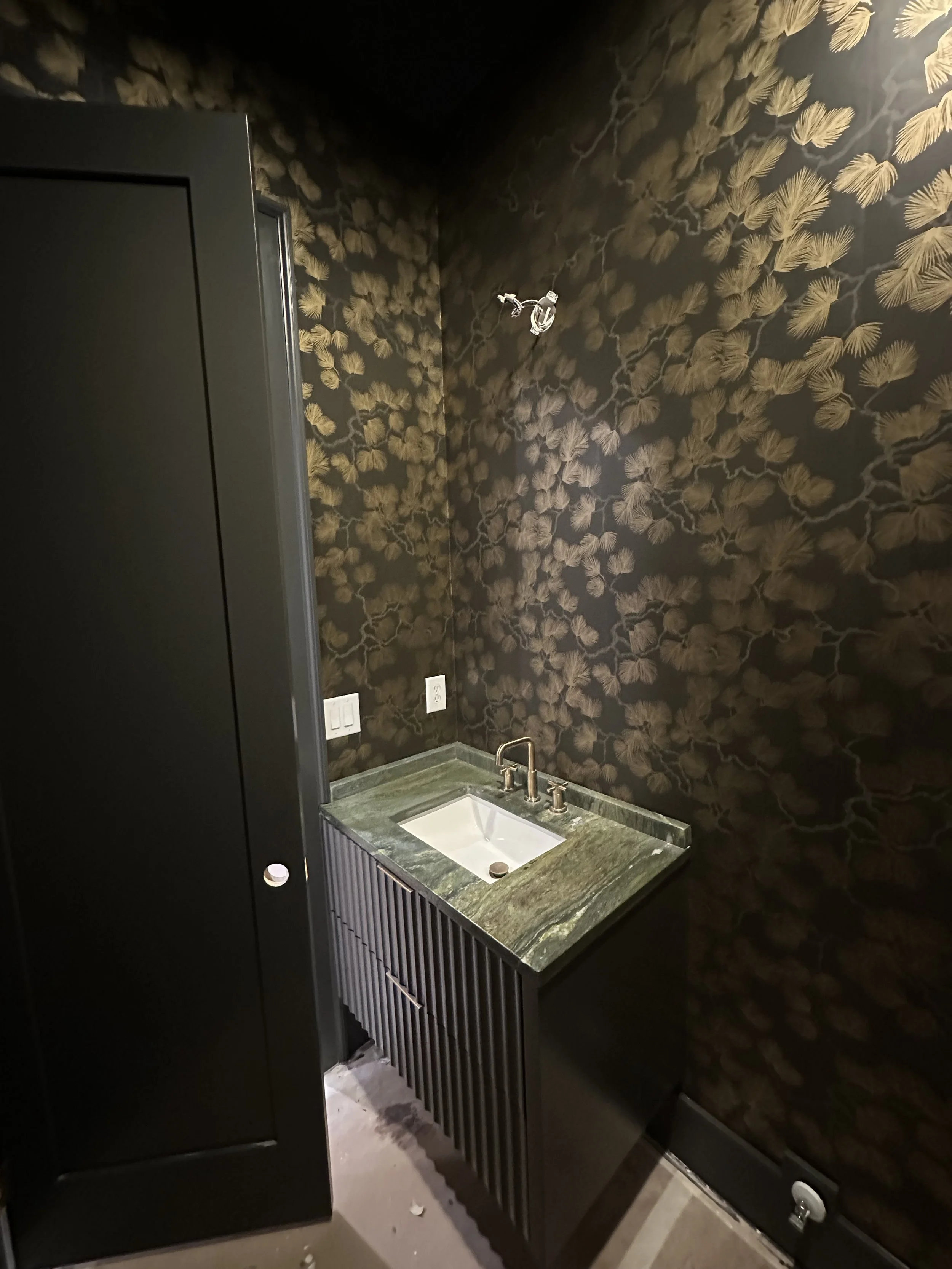 Small bathroom vanity with a green marble countertop and white sink, black cabinet with slatted front, dark wallpaper with gold leaf pattern, electrical outlets on the wall, and a wall-mounted faucet.