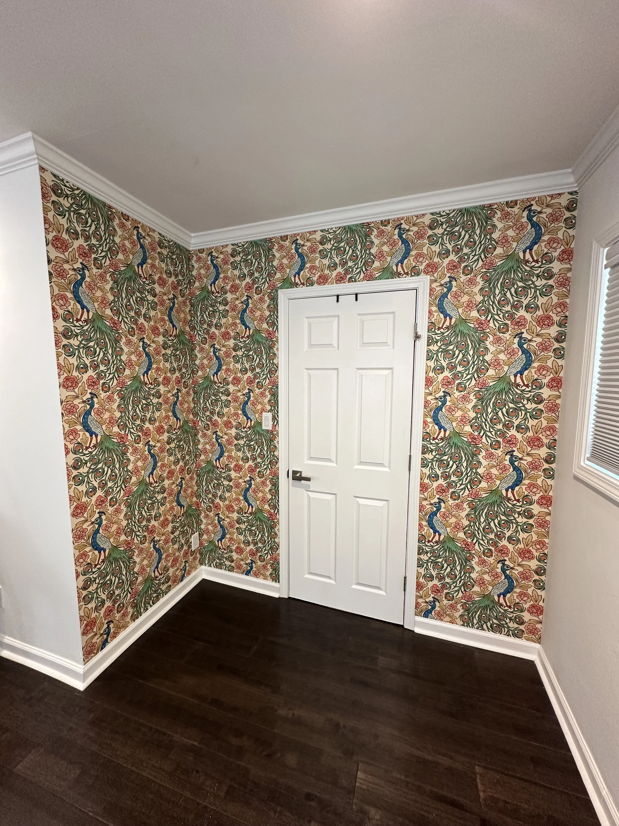 Room with peacock-themed wallpaper featuring multiple peacocks with blue and green feathers and red accents on a beige background, white door, dark hardwood floor, and window with white blinds.
