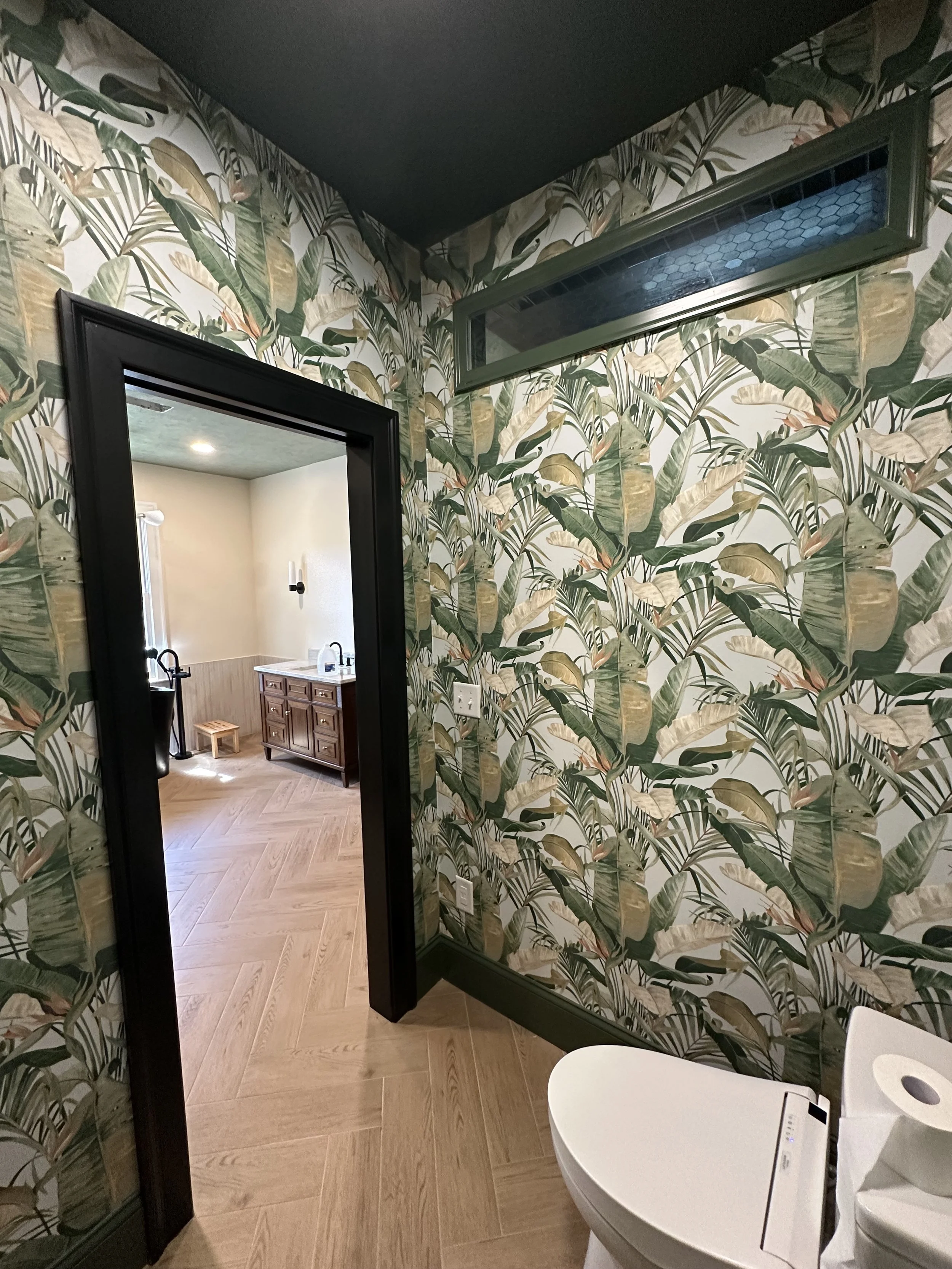Interior of a bathroom with tropical leaf wallpaper, a toilet on the right, and a doorway leading to a room with a wooden vanity and sink in the background.