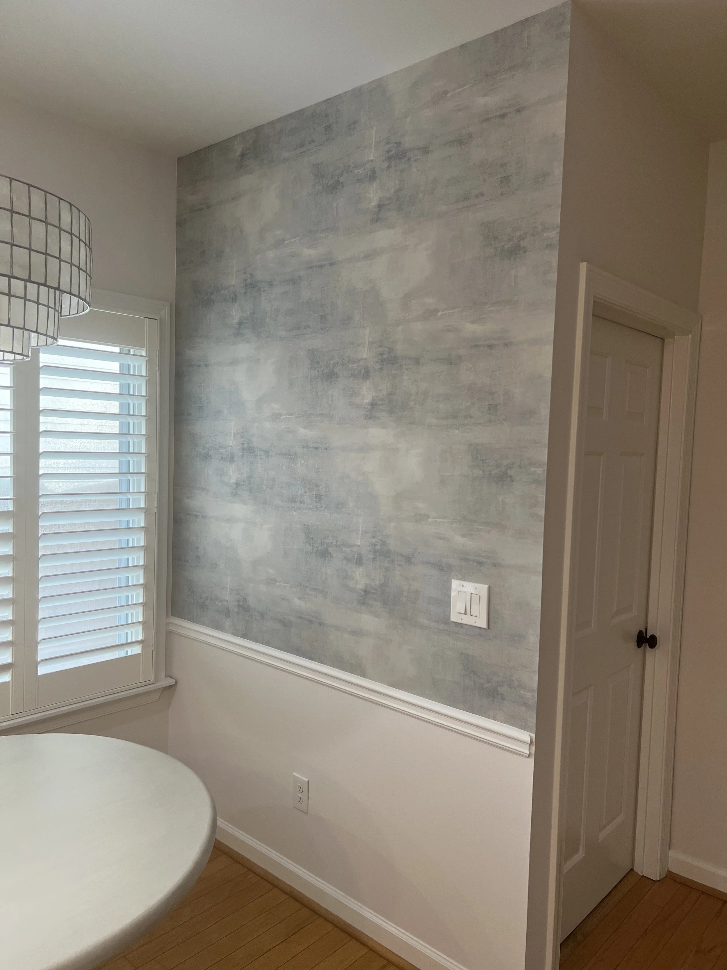 Interior corner of a room with a textured gray accent wall, a window with white plantation shutters, a white round table, and a closed white door.