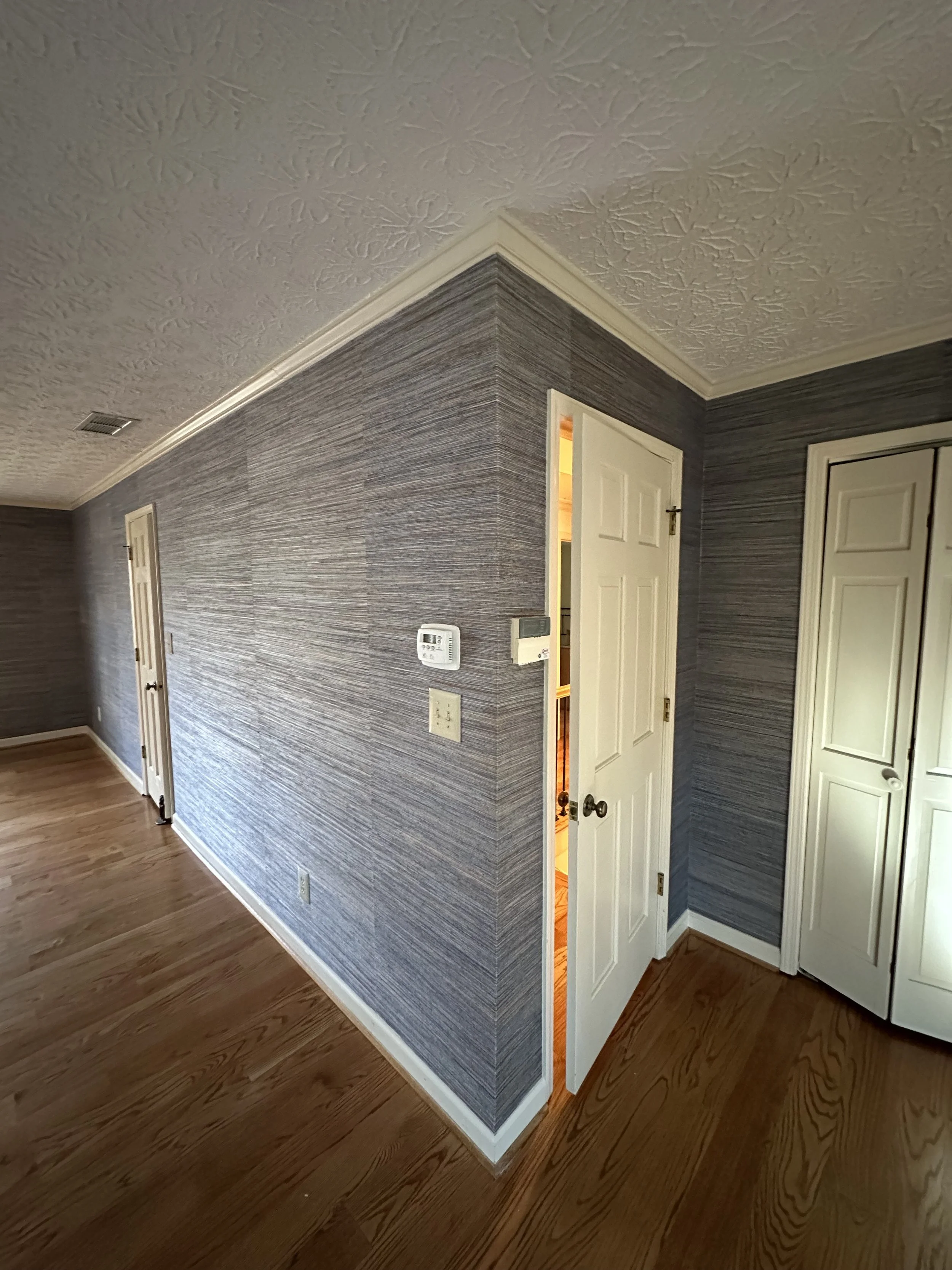 Room corner with textured gray wallpaper, white doors, light-colored wood flooring, and a visible thermostat on the wall.
