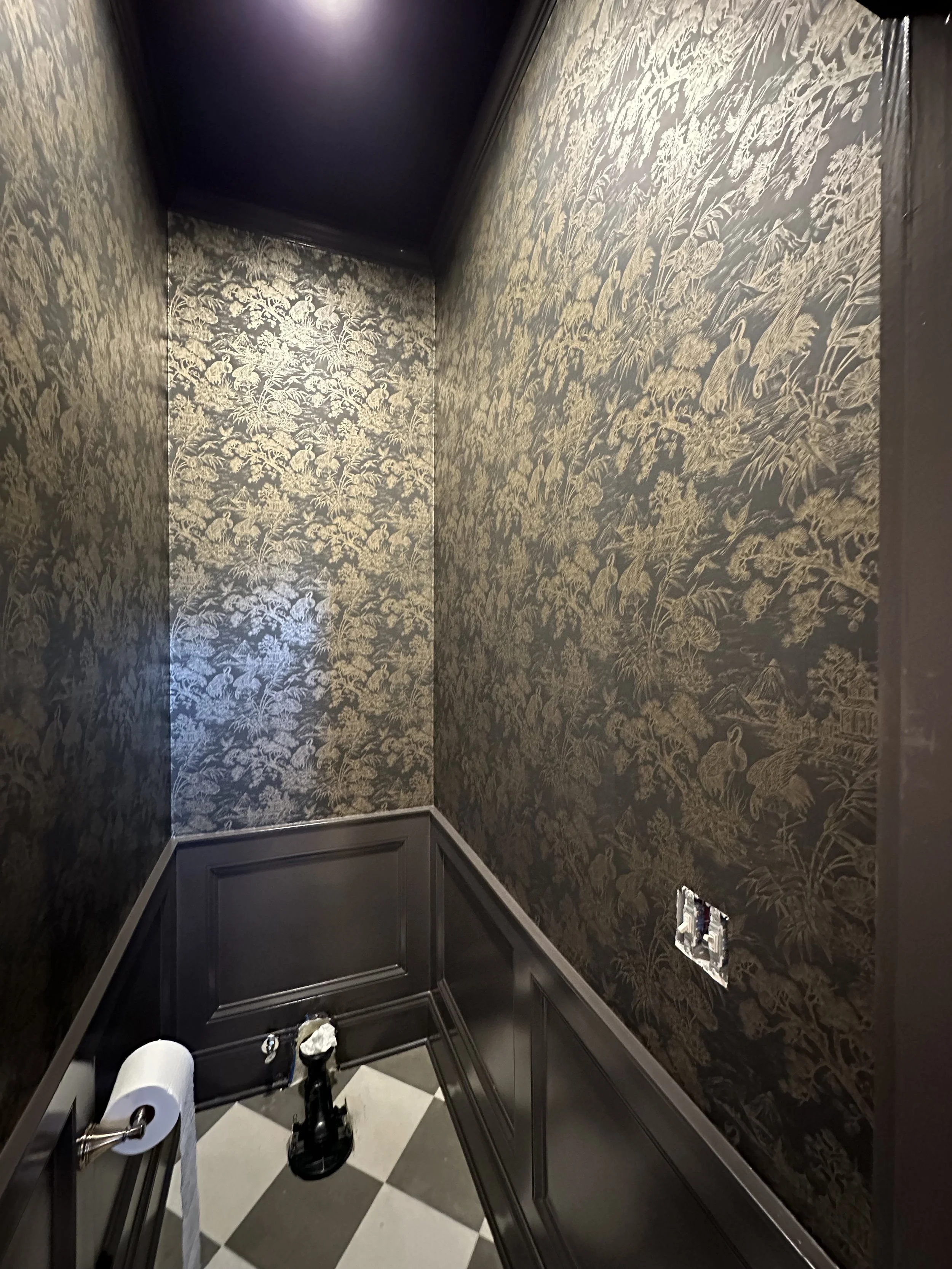 A small, decorative room with black wallpaper featuring a gold floral and bird pattern, black wainscoting, a checkered black and white tile floor, an electrical outlet without cover, and a toilet paper roll on a holder with a roll of toilet paper.