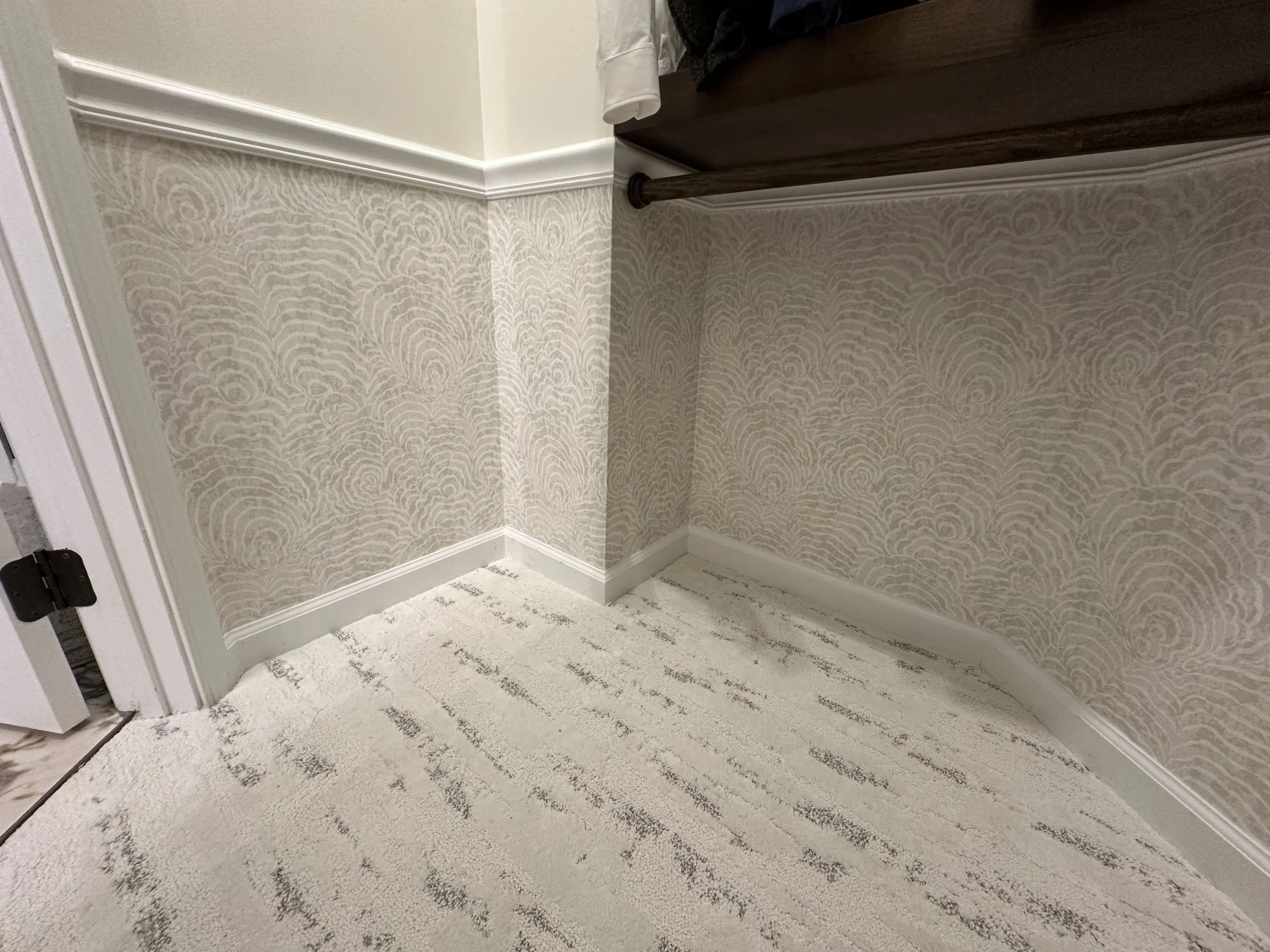 Interior corner of a room with patterned wallpaper on the walls, white baseboards, a beige carpeted floor, and part of a dark wooden piece of furniture.