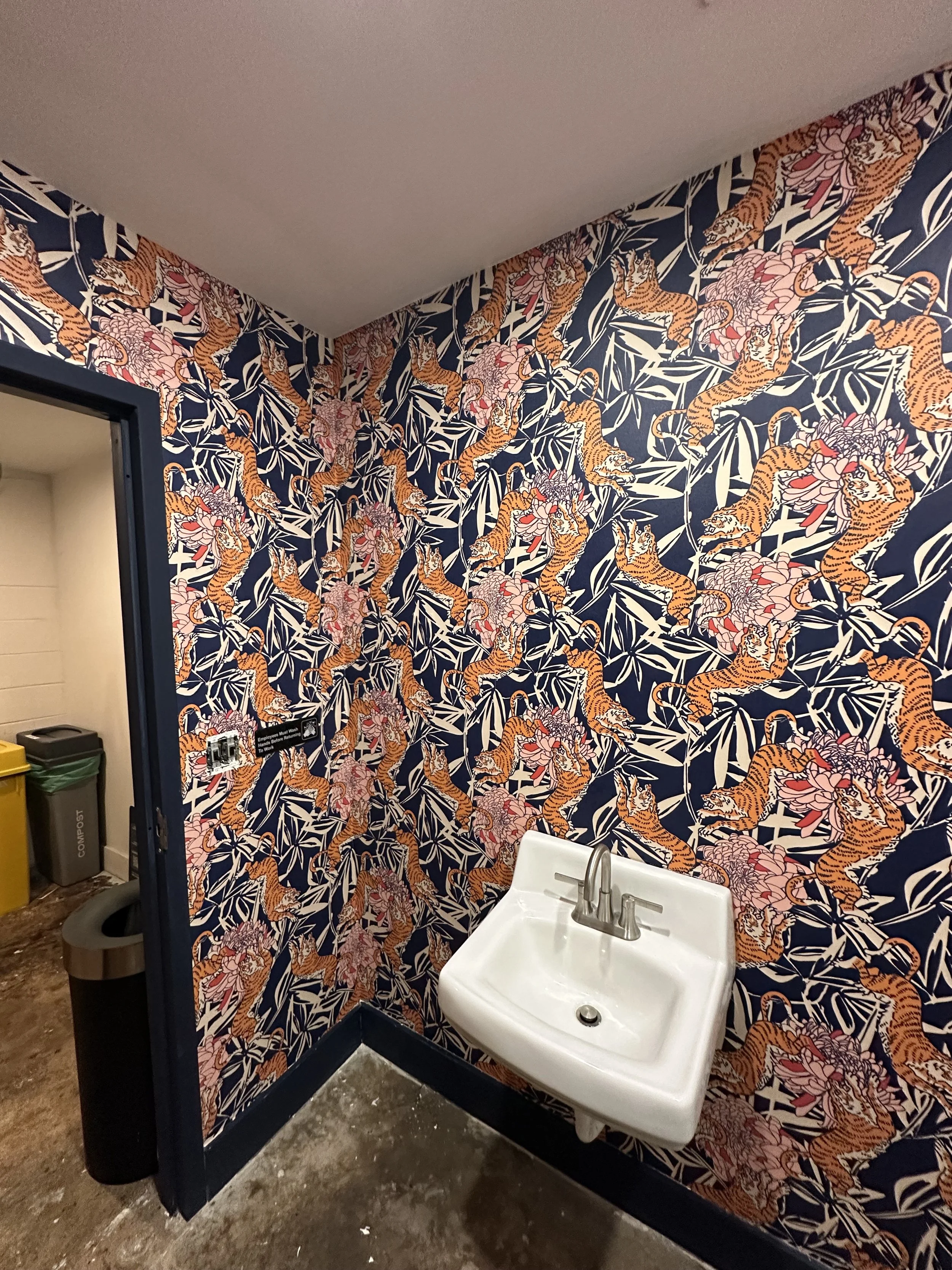 A corner of a bathroom with wallpaper decorated with orange tigers and pink flowers on a dark background, a white wall-mounted sink, and a black door frame leading to a room with trash cans.