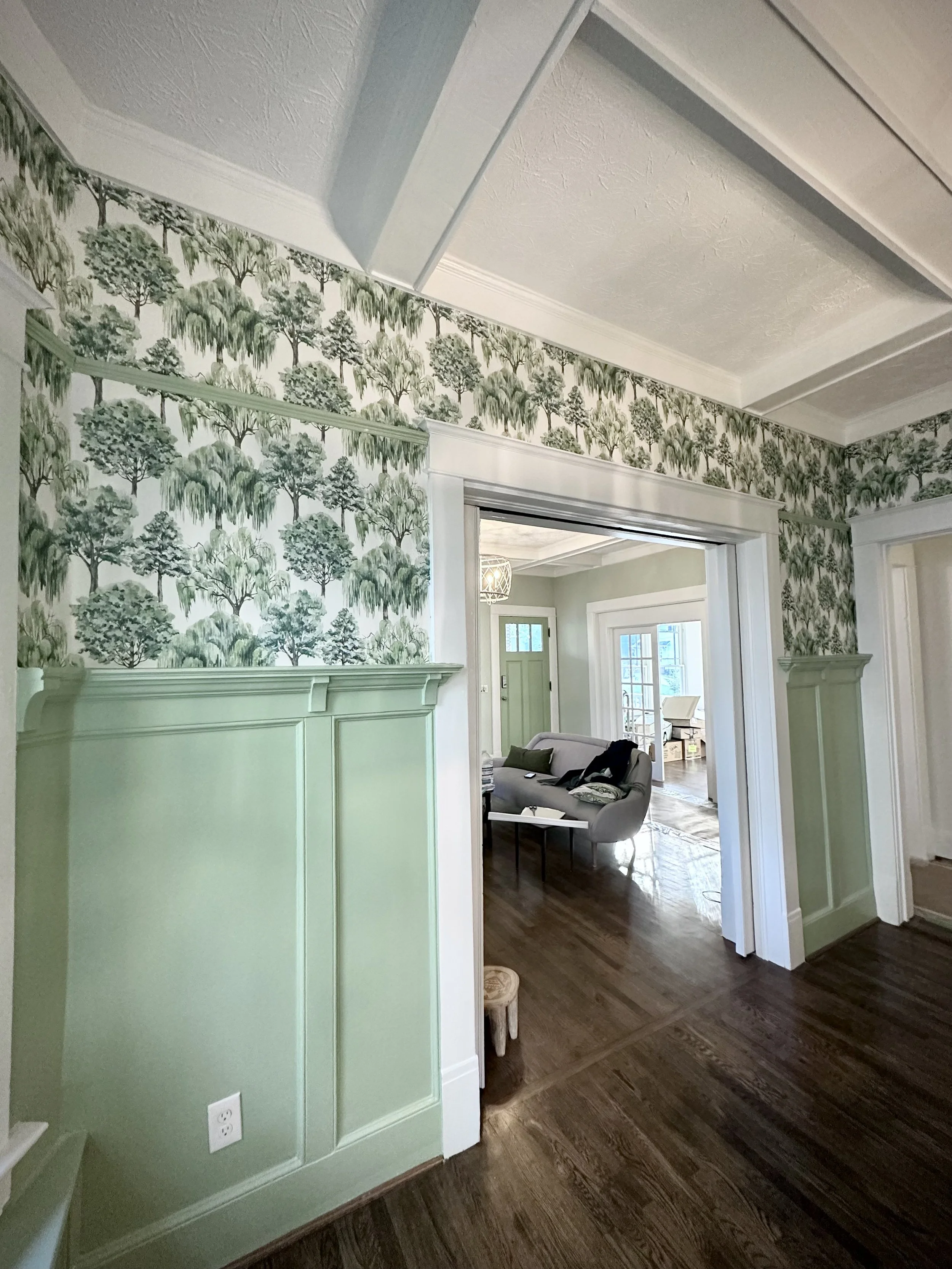 View of a room with green wallpaper featuring trees, a partial door frame, and a living area with a gray sofa, wooden floor, and glass doors leading outside.