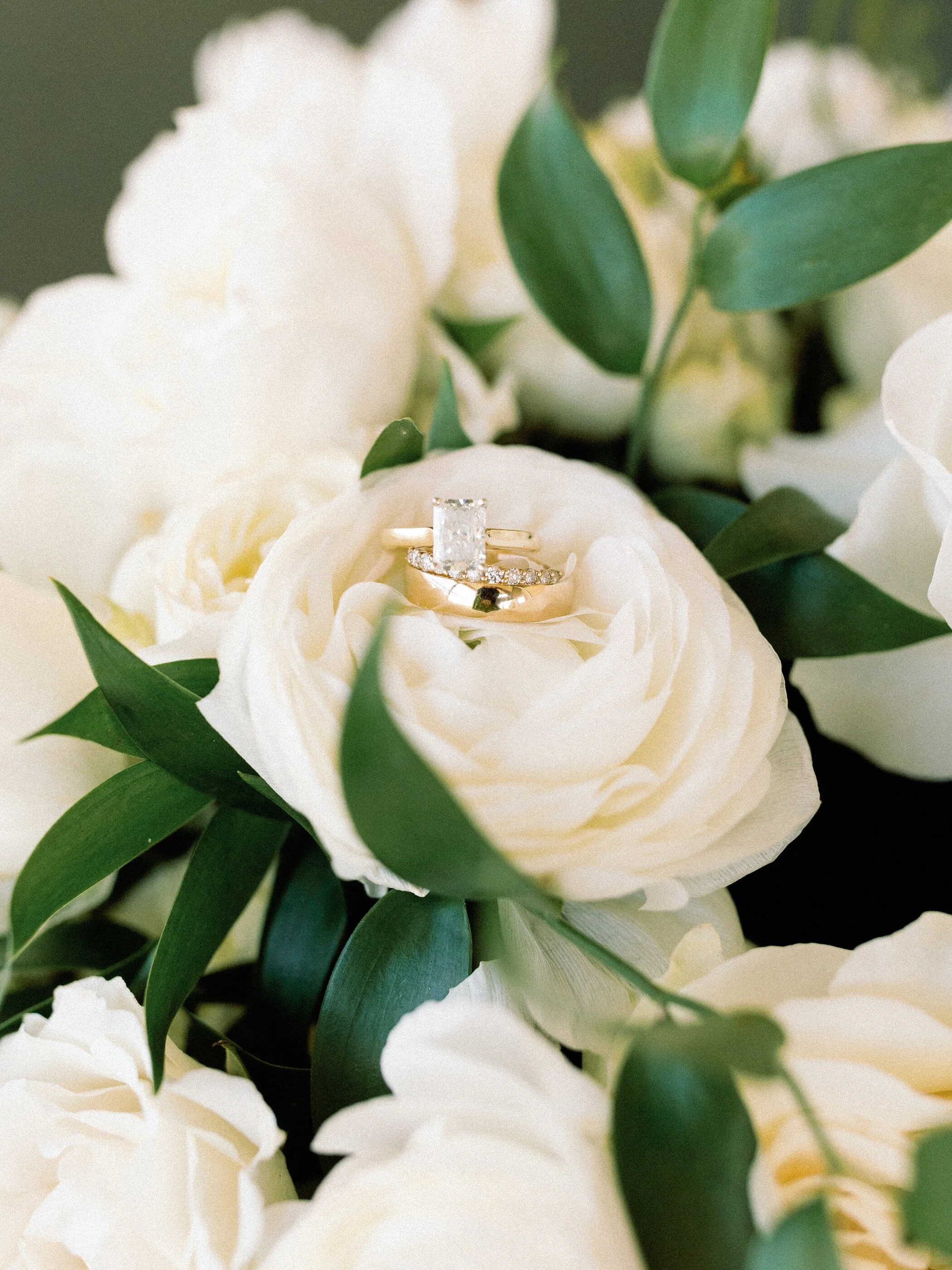 Gold engagement ring and wedding band on a white rose with green leaves.