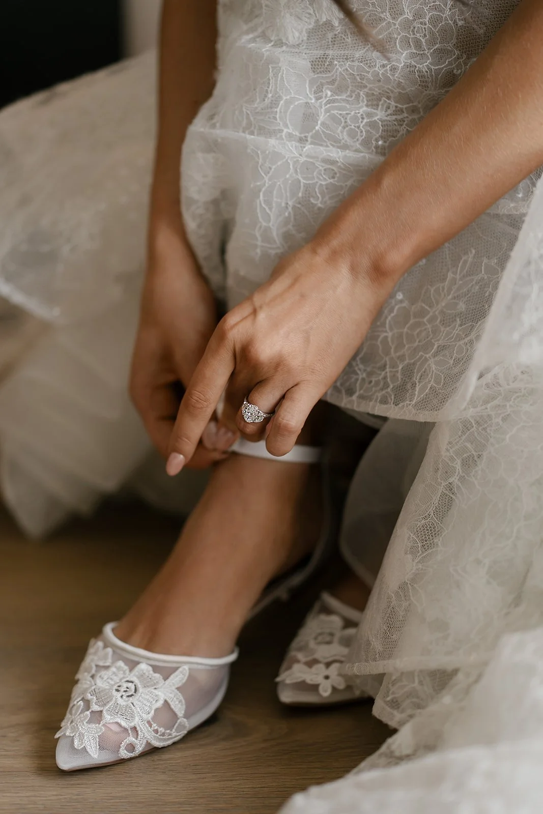Bride adjusting lace wedding shoes while wearing a lace dress and diamond ring.