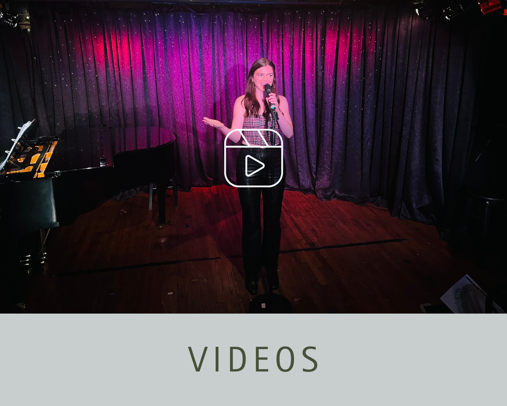 A woman standing on stage, holding a microphone, in front of a purple velvet curtain, with a grand piano to her left and pinkish stage lighting.