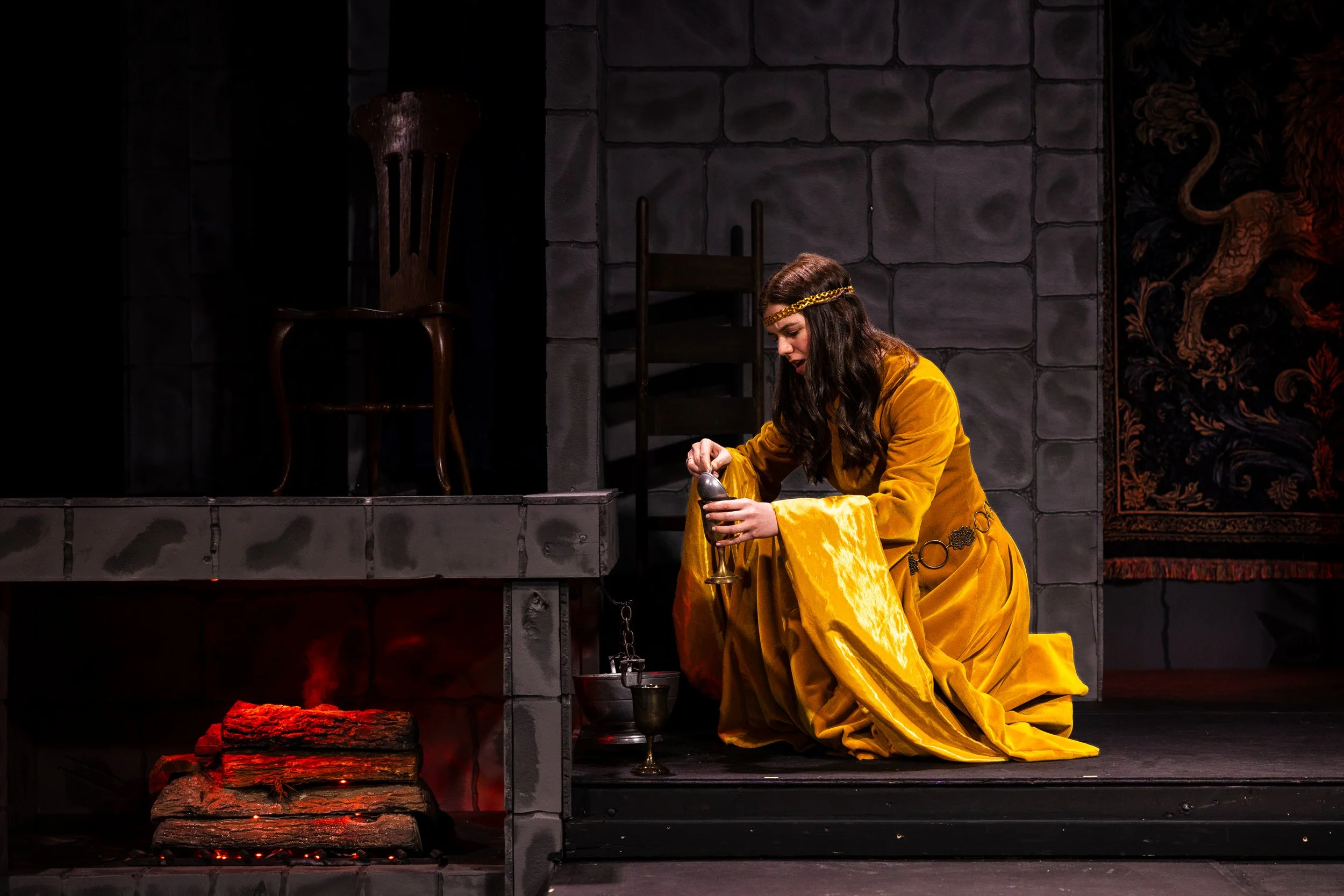 A woman dressed as a medieval princess or noblewoman in a yellow gown with long sleeves kneels on a stage, holding a chalice, in front of a painted stone wall backdrop. The stage includes a small fireplace with logs and red lighting, two chairs, and 