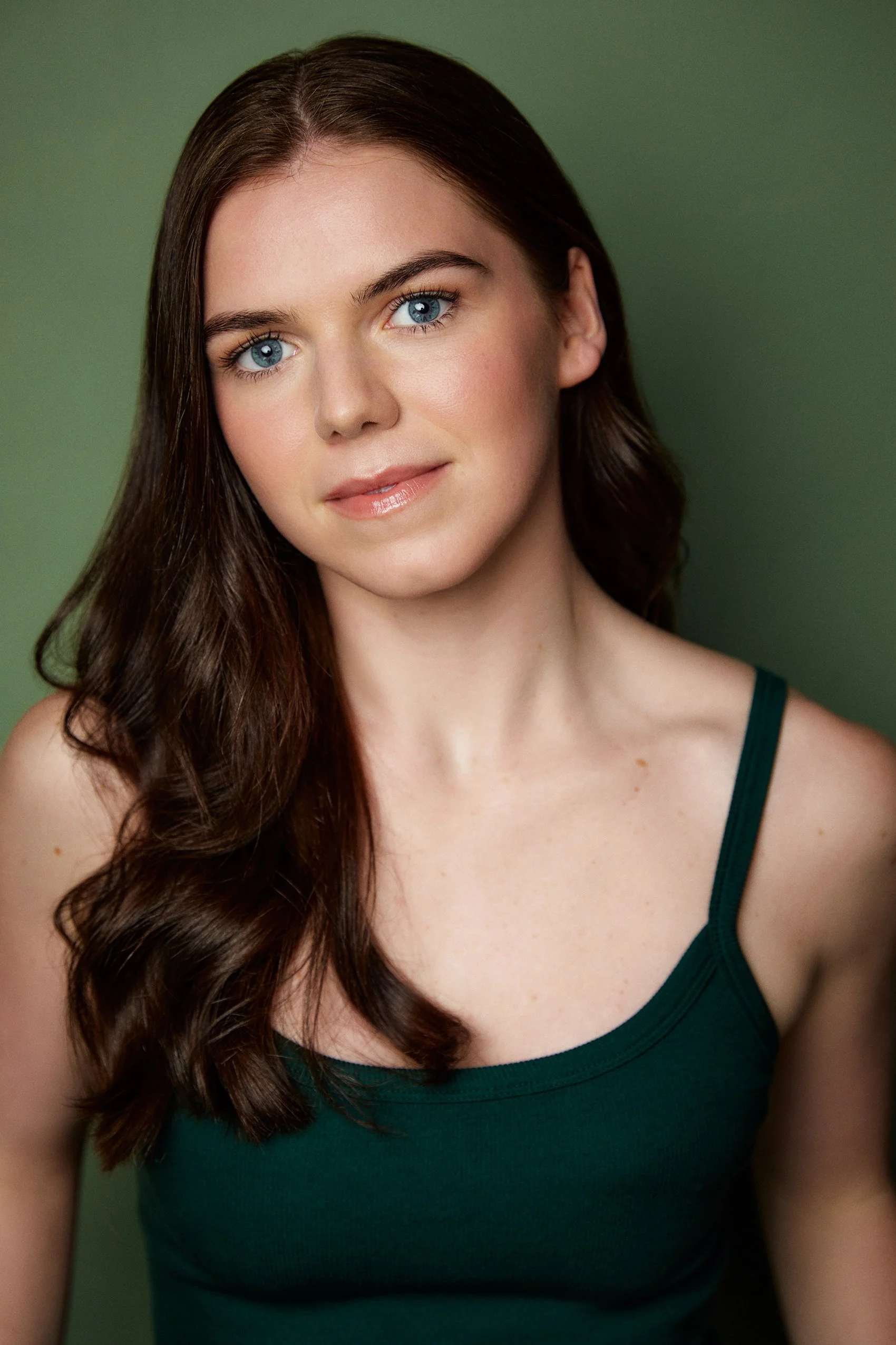 Close-up of a woman with long, wavy brown hair, blue eyes, wearing a dark green tank top against a green background.