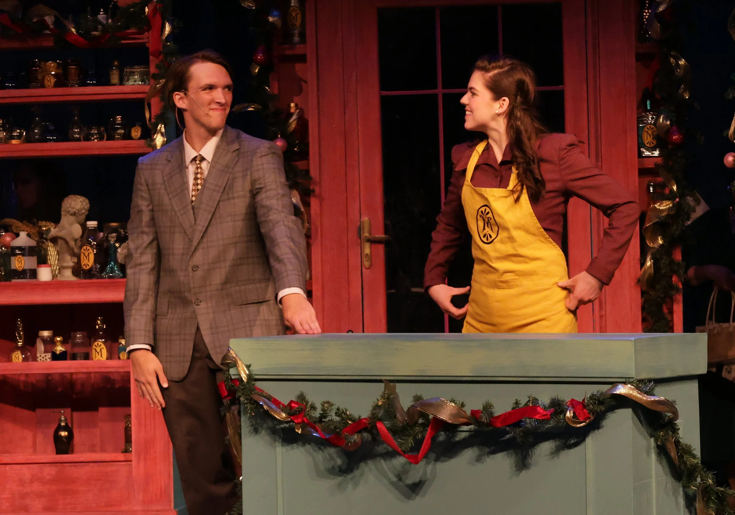 A man in a plaid suit and a woman in a yellow apron are standing behind a decorated table, smiling at each other in a festive setting with holiday decorations around them.