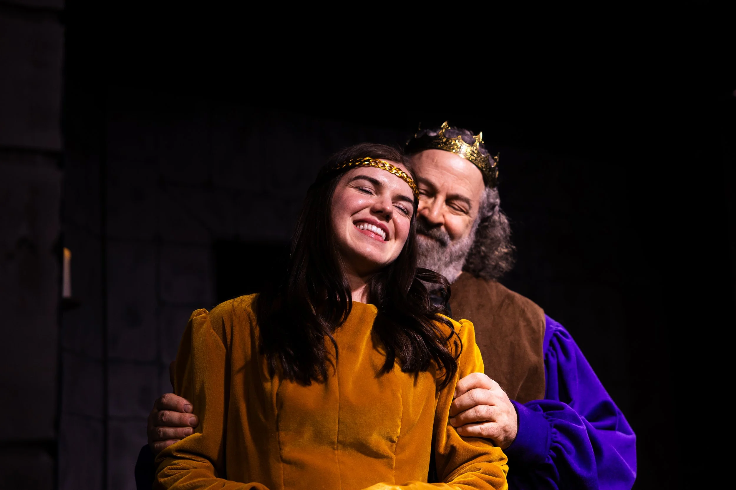 A young woman with long dark hair wearing a gold crown and a yellow dress standing next to an older man with a gray beard wearing a silver crown and a brown robe, both smiling with eyes closed, against a black background.