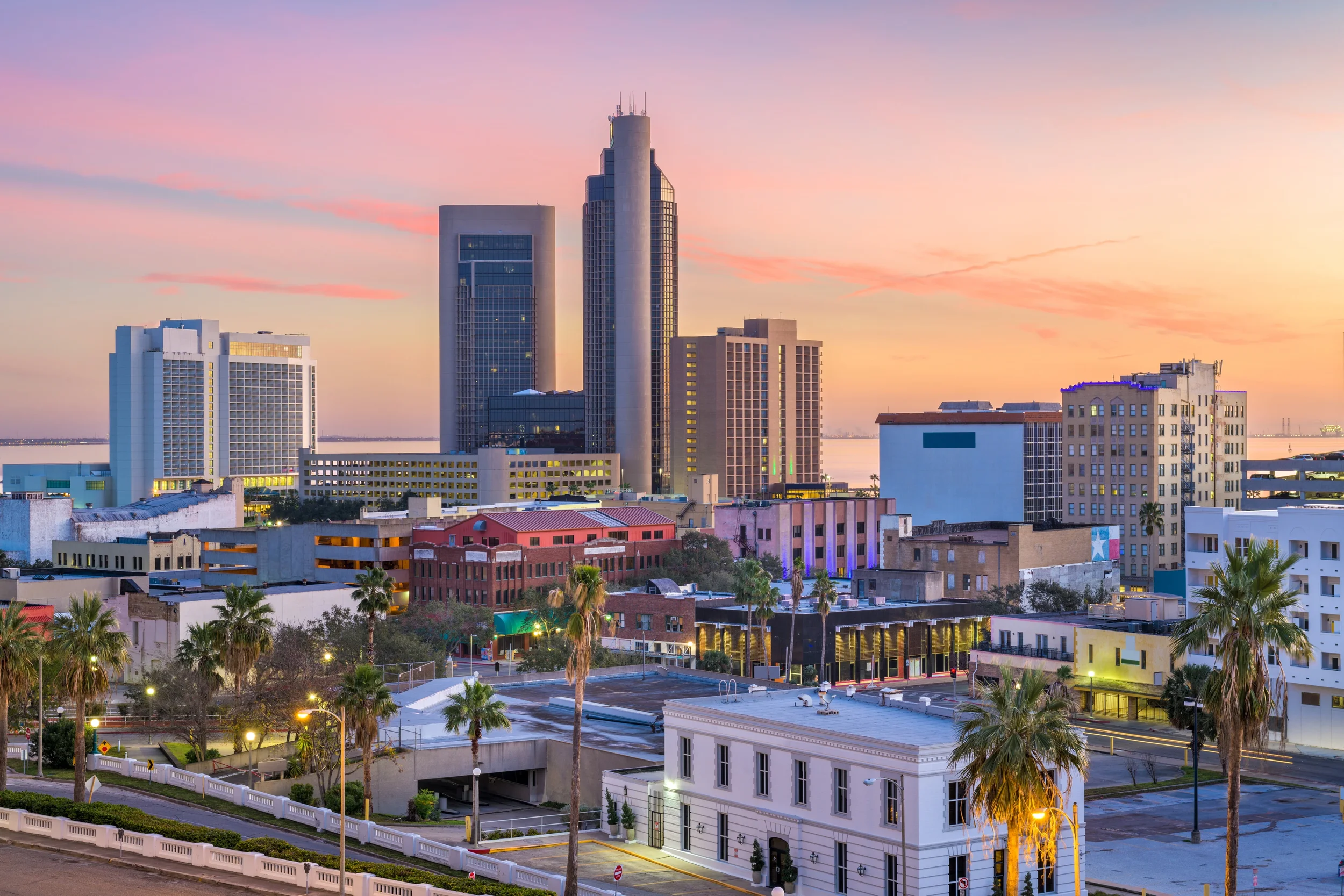 City skyline at sunset with tall modern skyscrapers, shorter buildings, palm trees, and city streets.