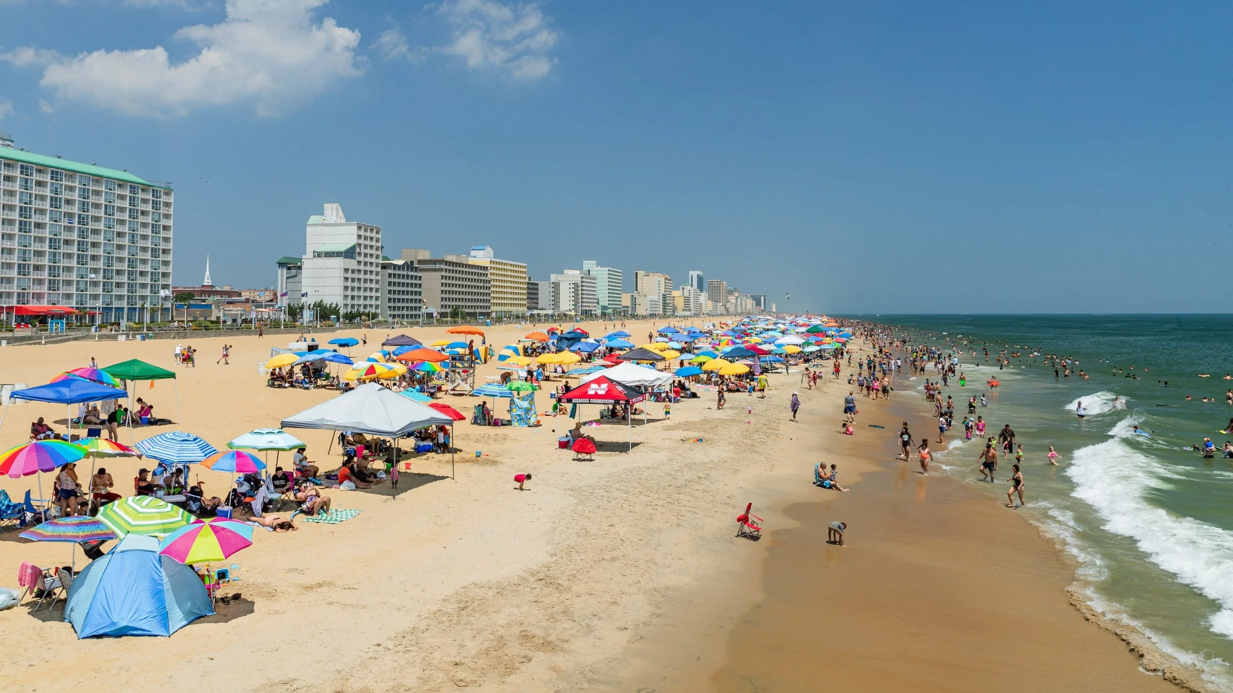  crowded beach with colorful umbrellas, people swimming, and high-rise buildings along the shore on a sunny day.