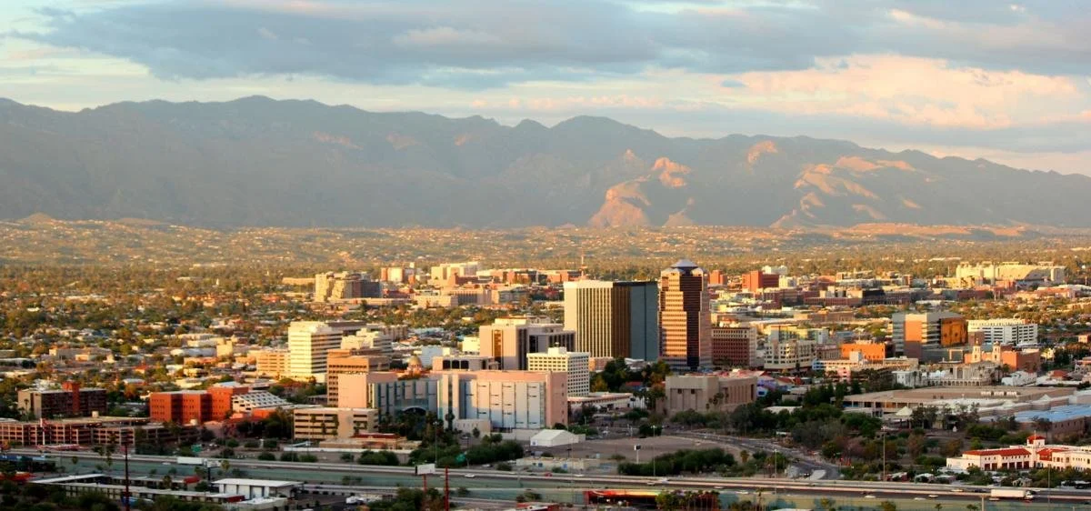 A city skyline with tall buildings in foreground and mountains in background, under a partly cloudy sky.