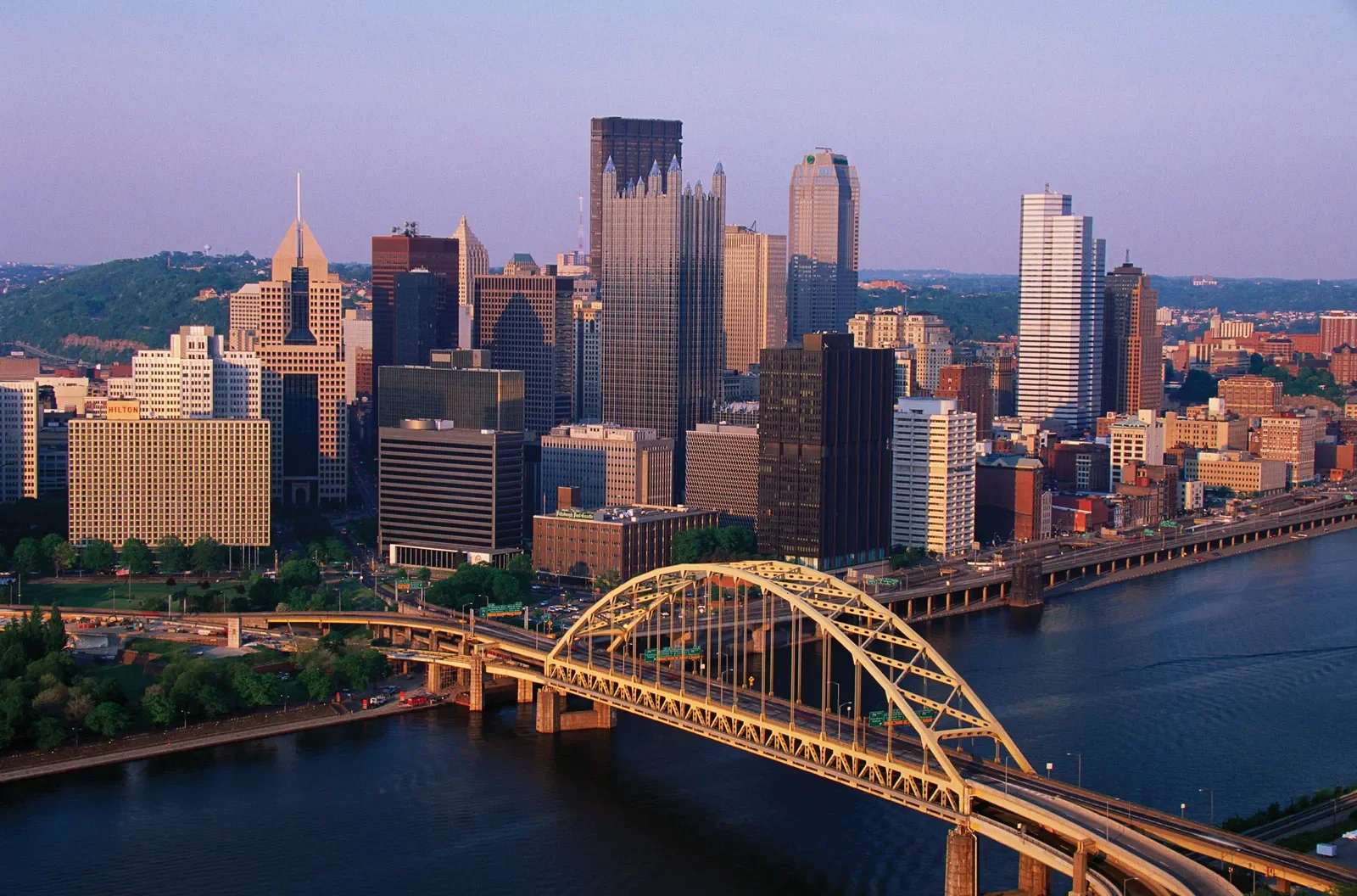 A skyline view of Pittsburgh, Pennsylvania, with high-rise buildings and the yellow bridge over the river at sunset.