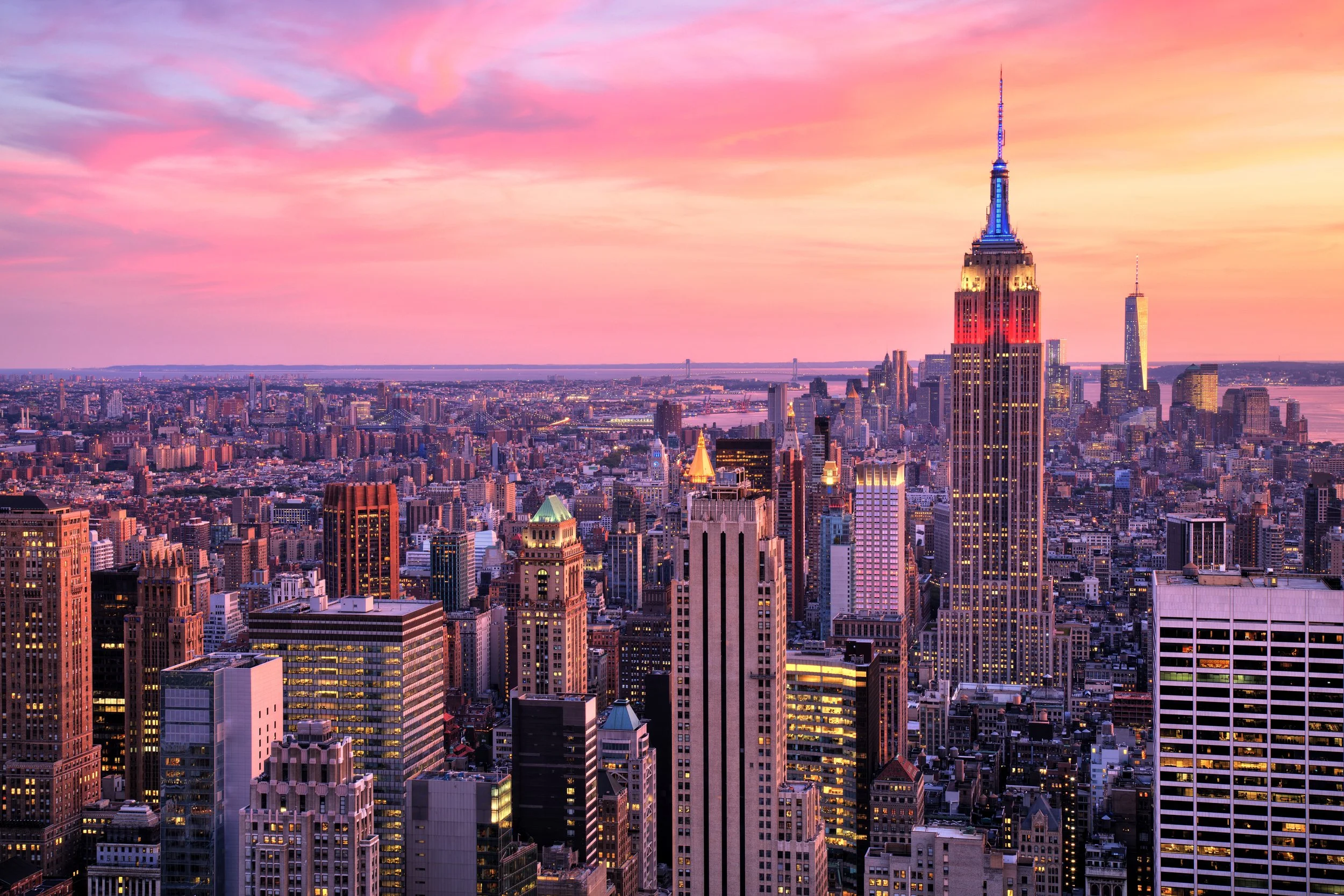 A city skyline at sunset featuring the Empire State Building illuminated with pink and blue lights, surrounded by other tall skyscrapers and buildings.