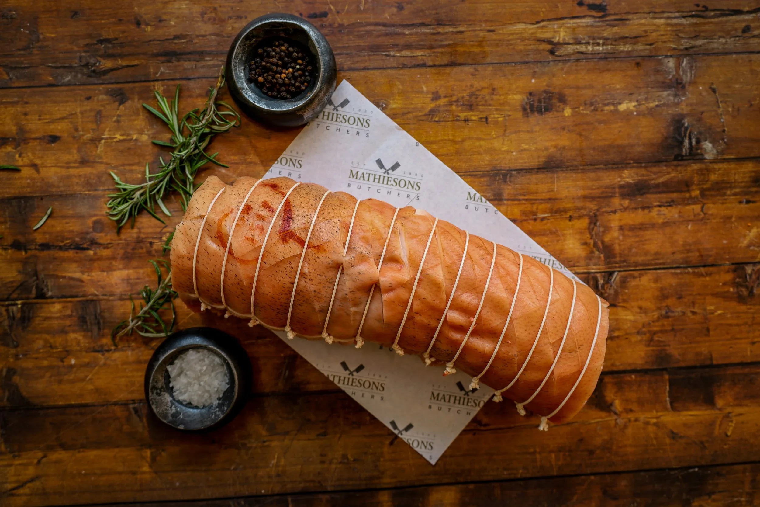 Raw tied pork tenderloin on parchment paper with rosemary, black pepper, and salt on a wooden surface.