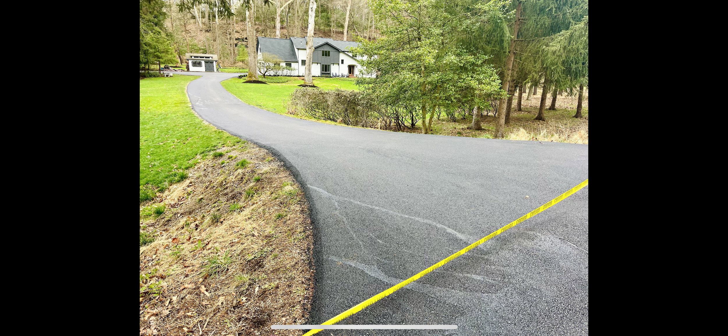 A winding paved driveway leading to a modern white house surrounded by lush green grass and trees, with yellow caution tape at the edge of the driveway indicating a possible investigation or hazard.