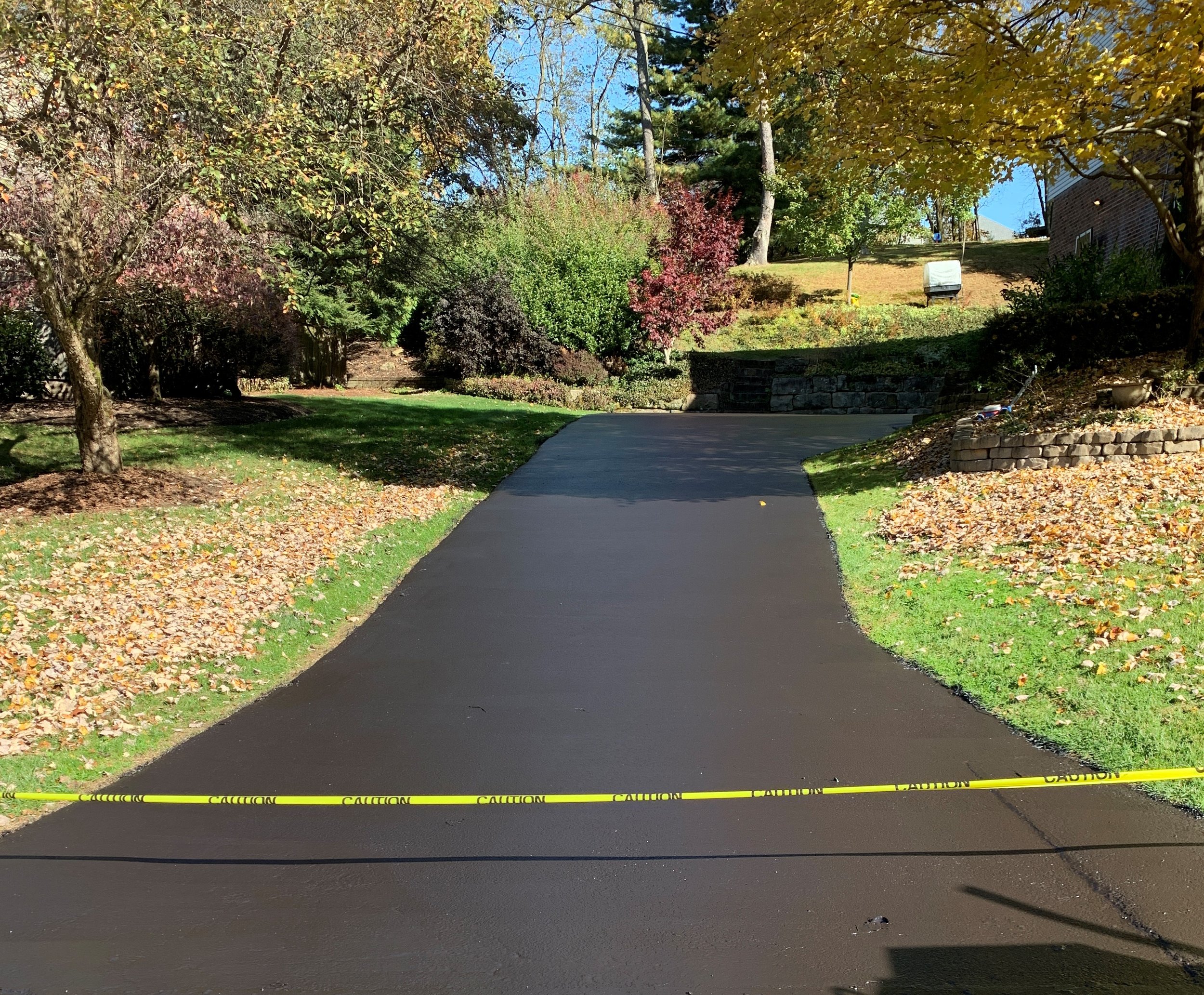 A paved road in a residential neighborhood is blocked off with yellow caution tape. There are trees with fall foliage, a grassy slope, a brick building, and a mailbox in the background.