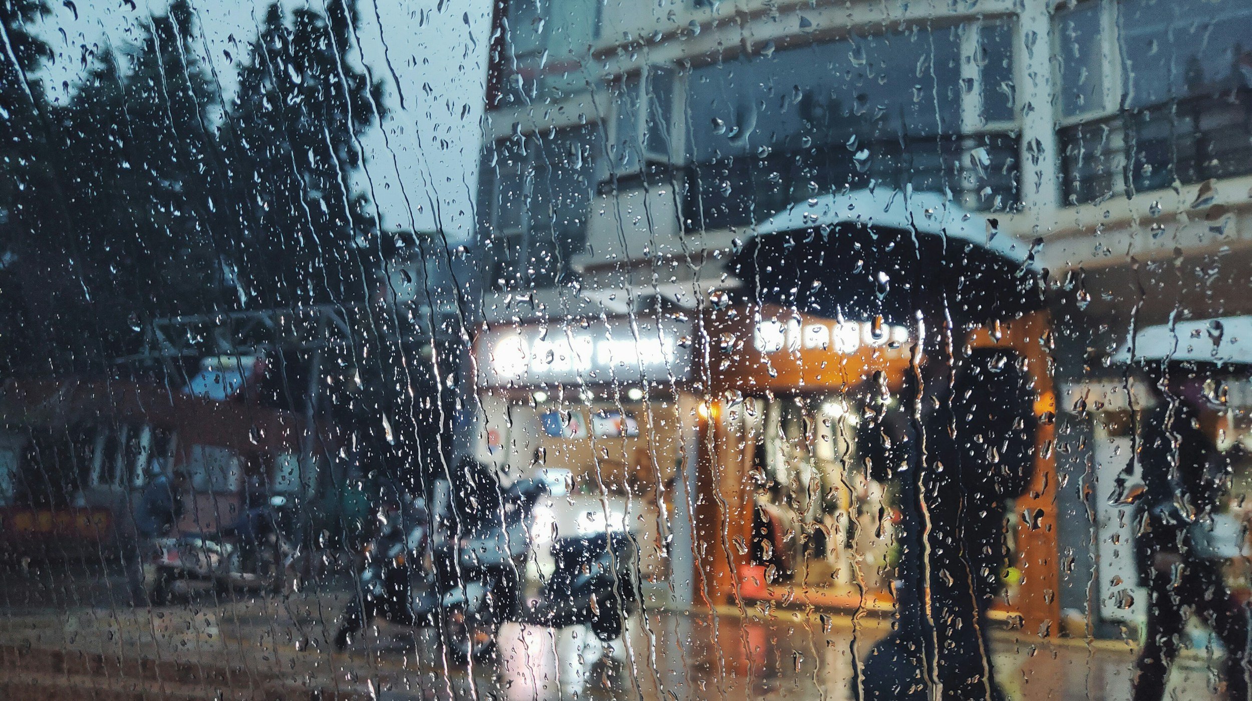 Rainy street view through a wet window, blurred figures with umbrellas, buildings in the background.