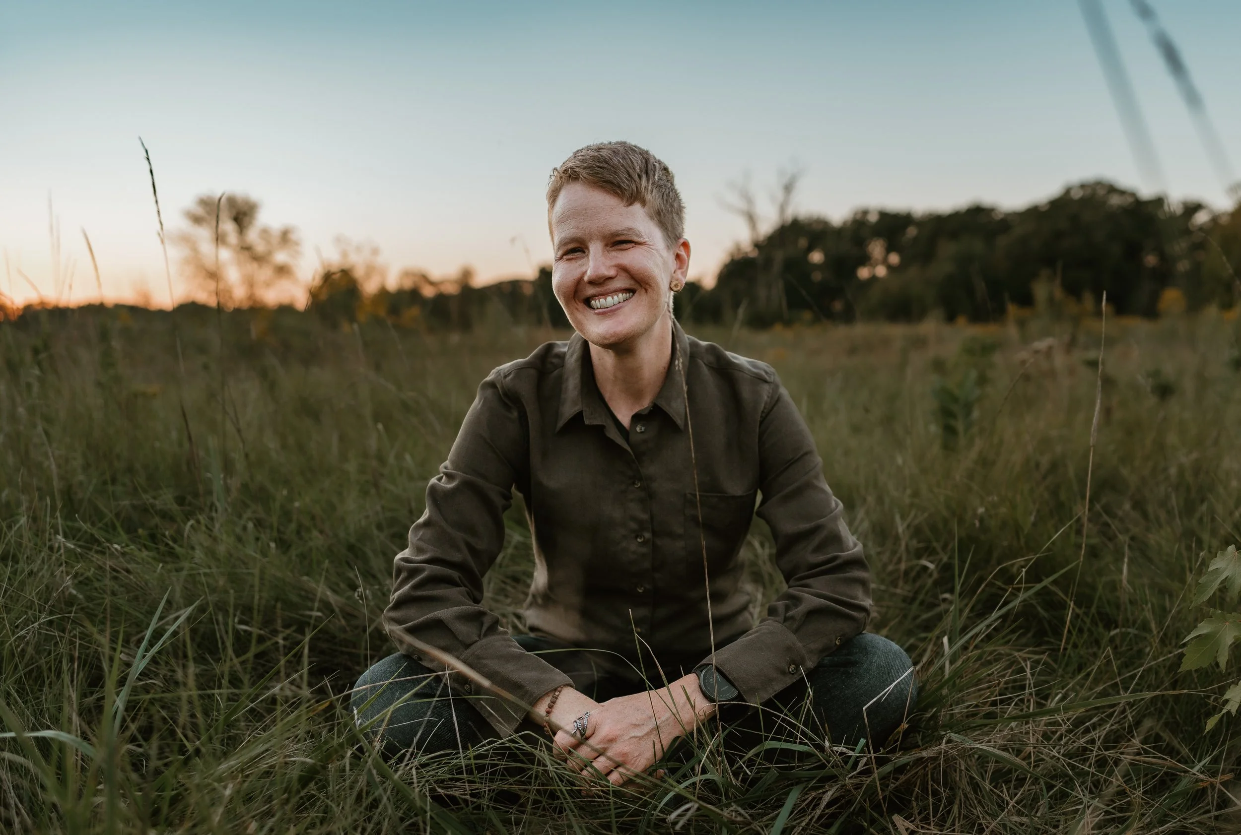 Corinne wearing a dark green button-up, seated cross-legged in a prairie with a wide smile